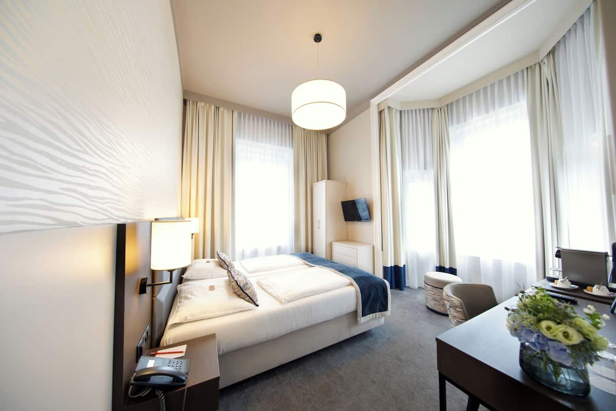 Habitación de hotel con cama, mesita de noche, lámpara, teléfono, televisión, escritorio, silla y un florero con flores.