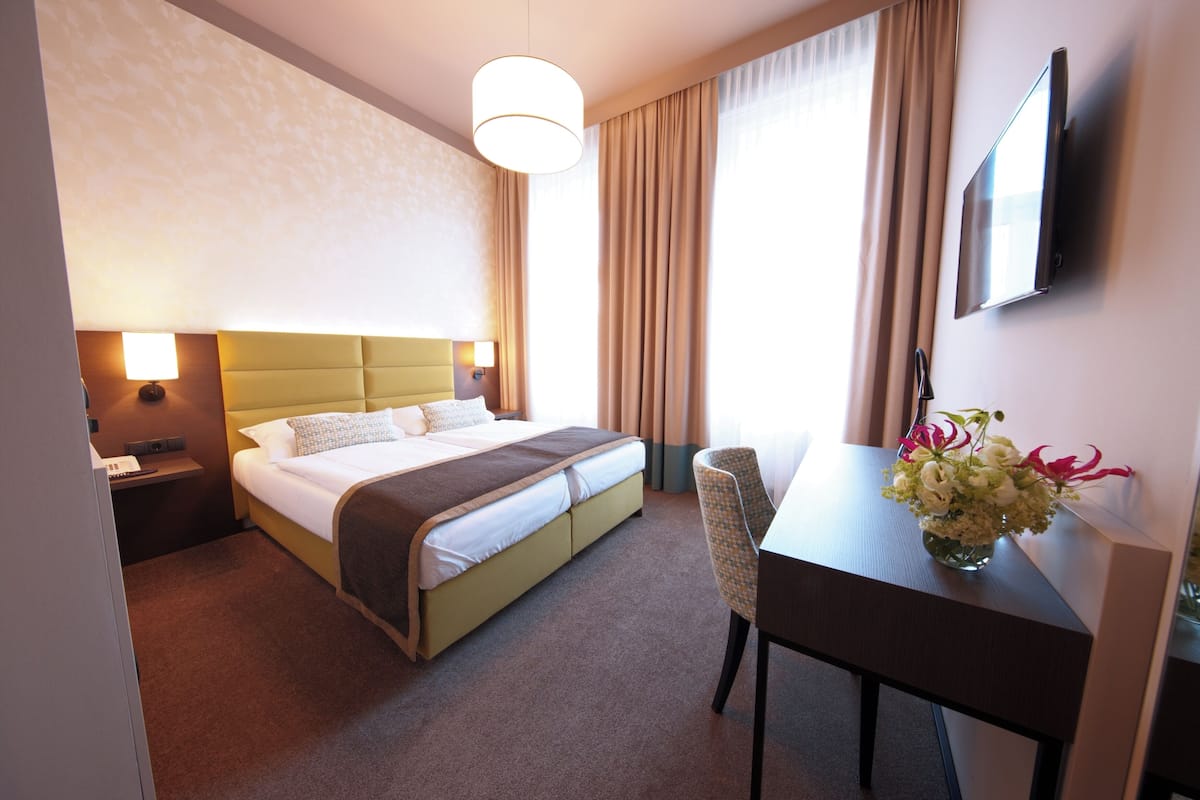 Habitación de hotel con cama, mesita de noche, lámpara, escritorio, silla y televisor montado en la pared.