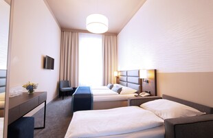Quarto de hotel com duas camas, uma escrivaninha, uma cadeira, televisão e uma janela grande com cortinas.