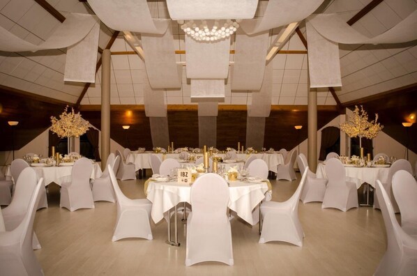 A banquet hall with round tables, white chairs, and chandeliers.