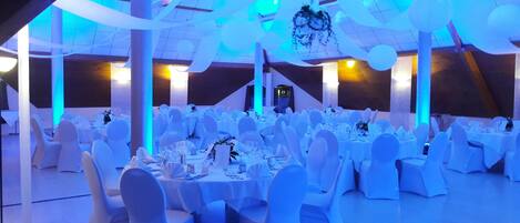 A banquet hall with round tables and white chairs, illuminated by blue lighting.