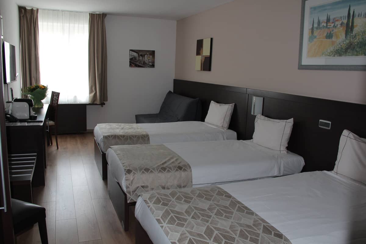 Hotellihuone, jossa on kolme sänkyä, työpöytä tuolilla ja ikkuna, jossa on verhot.
