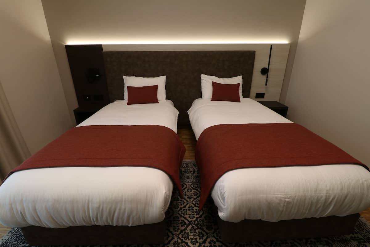 Hotellihuoneessa on kaksi sänkyä, joissa on valkoiset lakanat ja punaiset koristepaidat. Huoneessa on moderni sängynpääty ja kuviollinen matto.