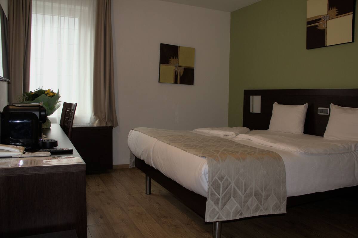 Hotellihuone, jossa on sänky, työpöytä, jossa on tietokone, tuoli ja ikkuna, jossa on verhot.