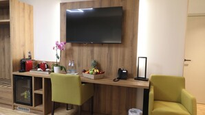 Zona de recepción de un hotel con un mostrador de madera, dos sillas verdes, un televisor, una cesta de frutas, una máquina de café, un teléfono y un refrigerador pequeño.