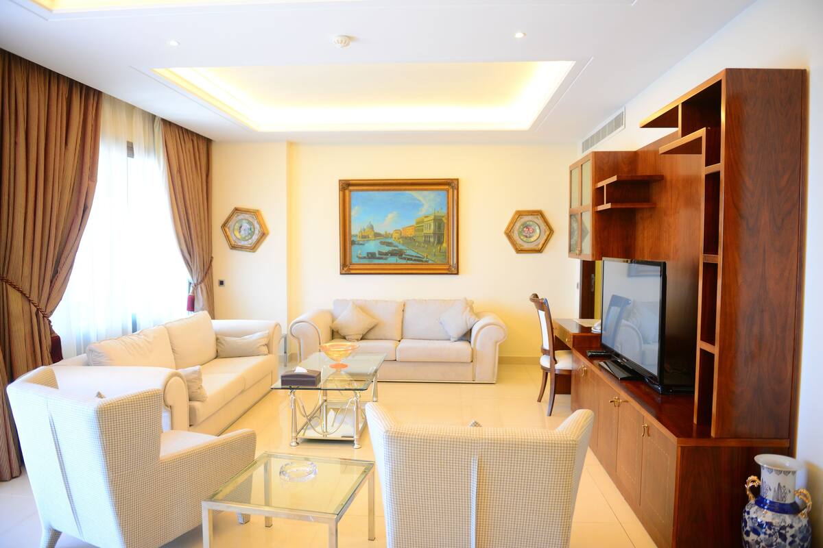 A living room with a sofa, armchairs, a glass coffee table, a TV on a wooden cabinet, and a painting on the wall.