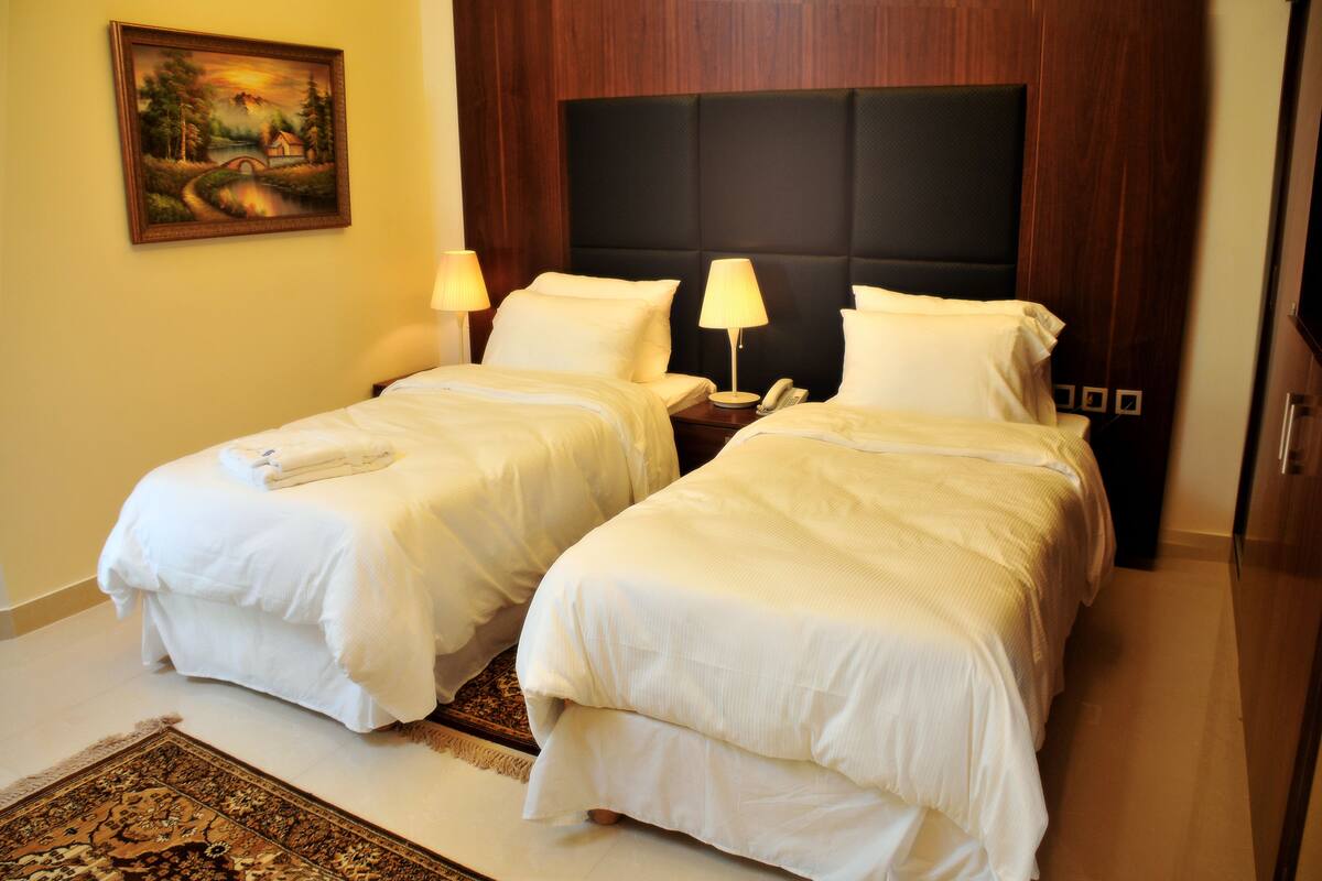 A hotel room with two single beds, a nightstand with a lamp, a painting on the wall, and a wooden headboard.