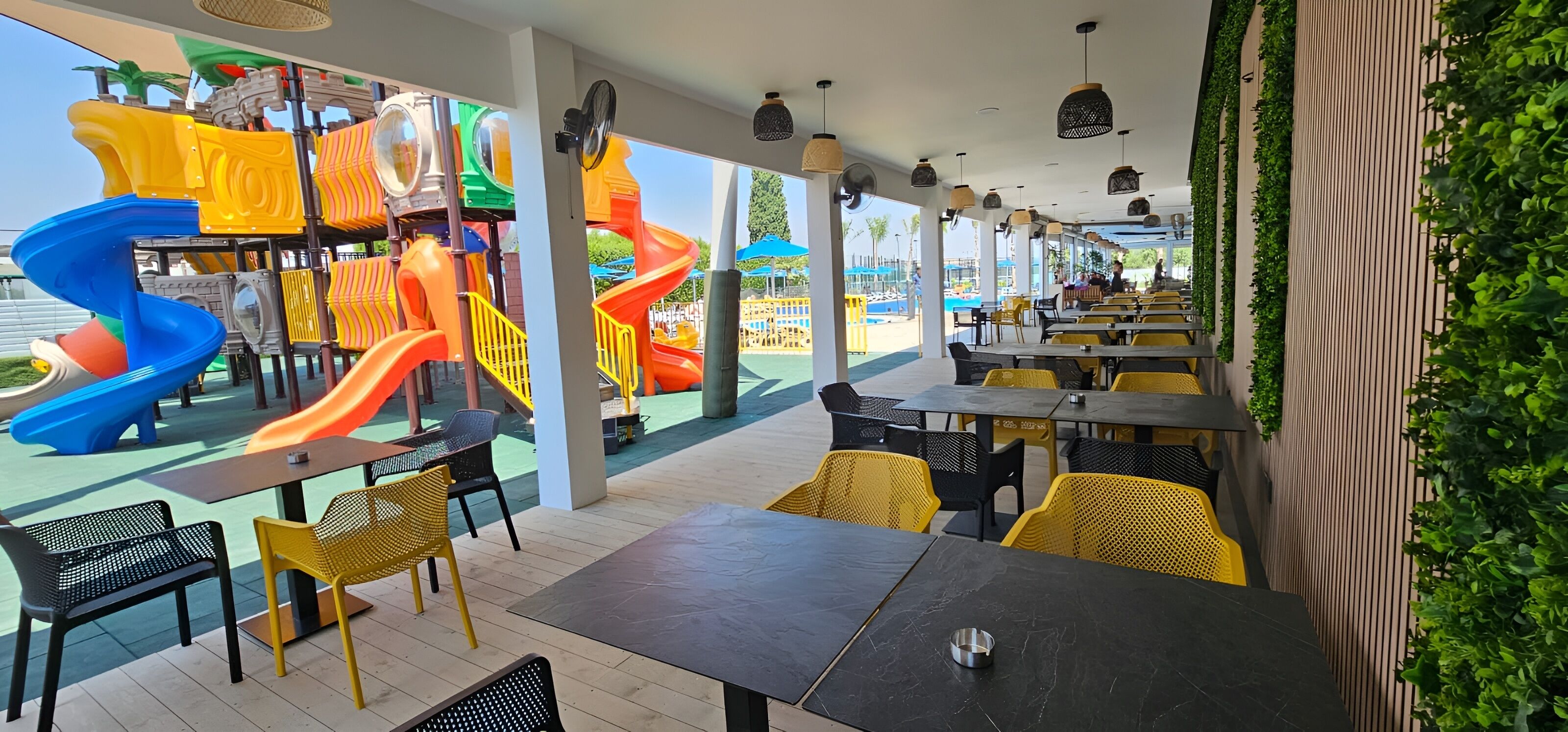 An outdoor dining area with tables and chairs, a children's play area with slides, and a view of a pool.