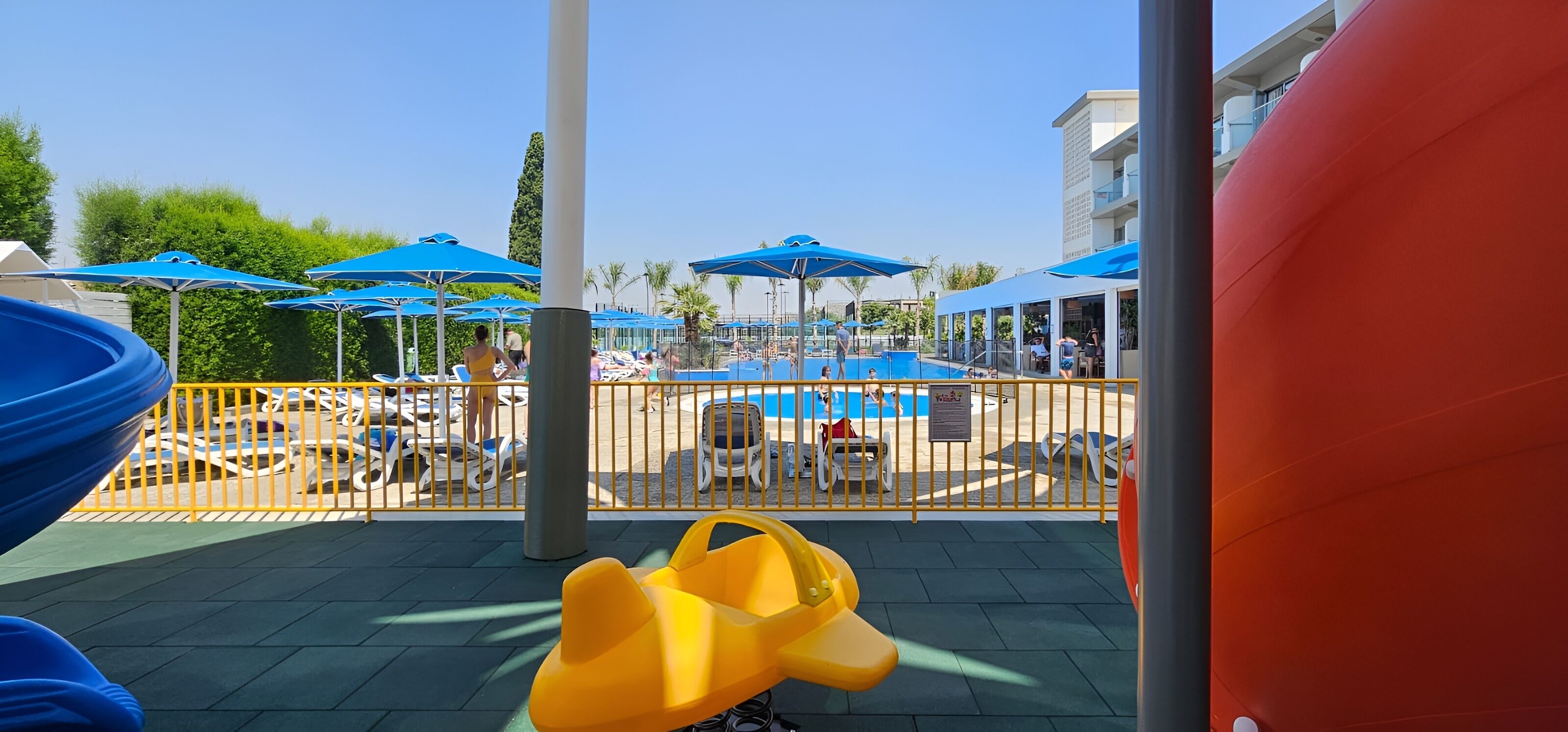 Zona de piscina con sombrillas, sillas de descanso y un vehículo para niños amarillo.
