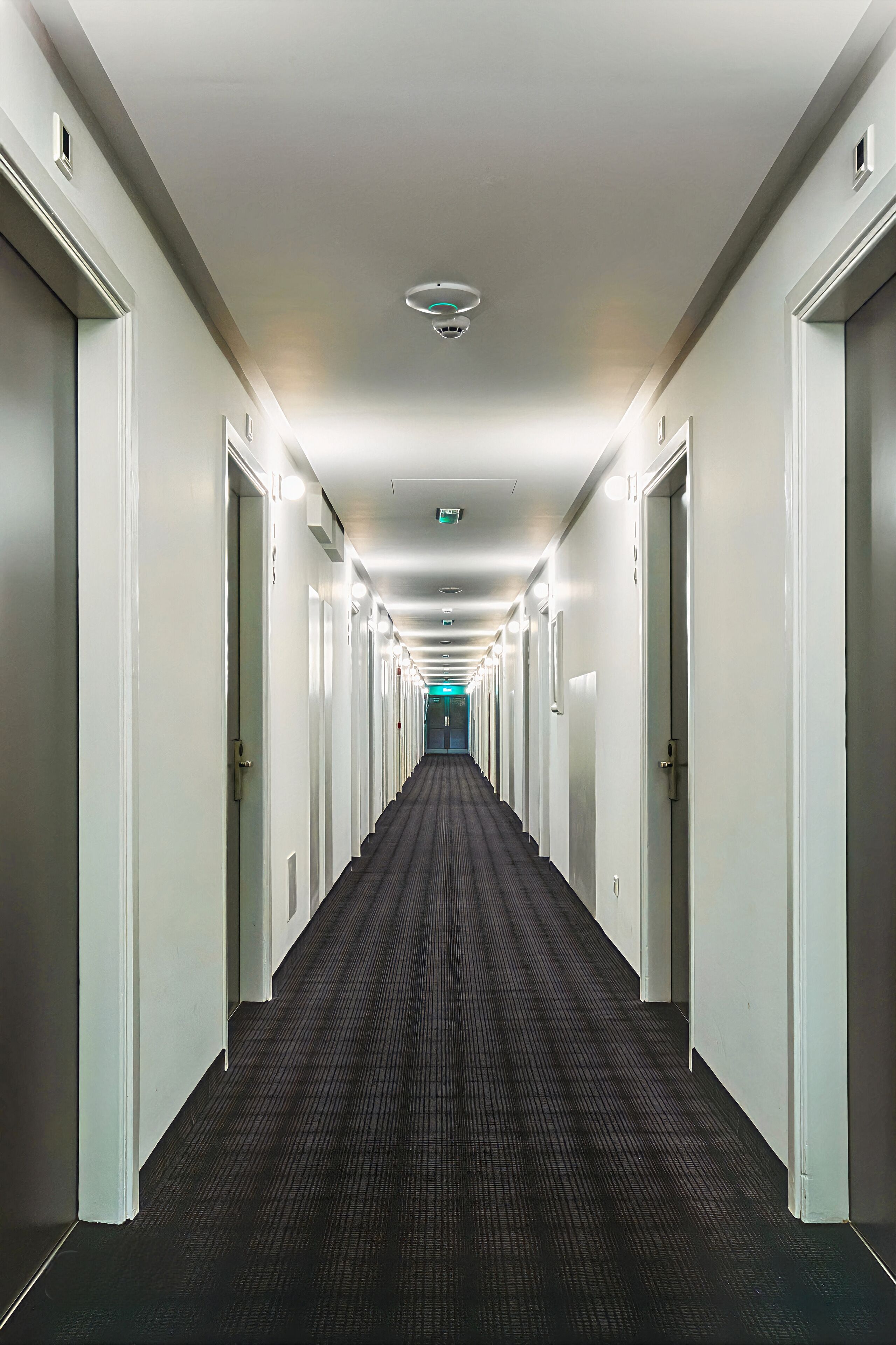 A long, narrow hotel corridor with doors on both sides, a patterned carpet, and a ceiling light fixture.