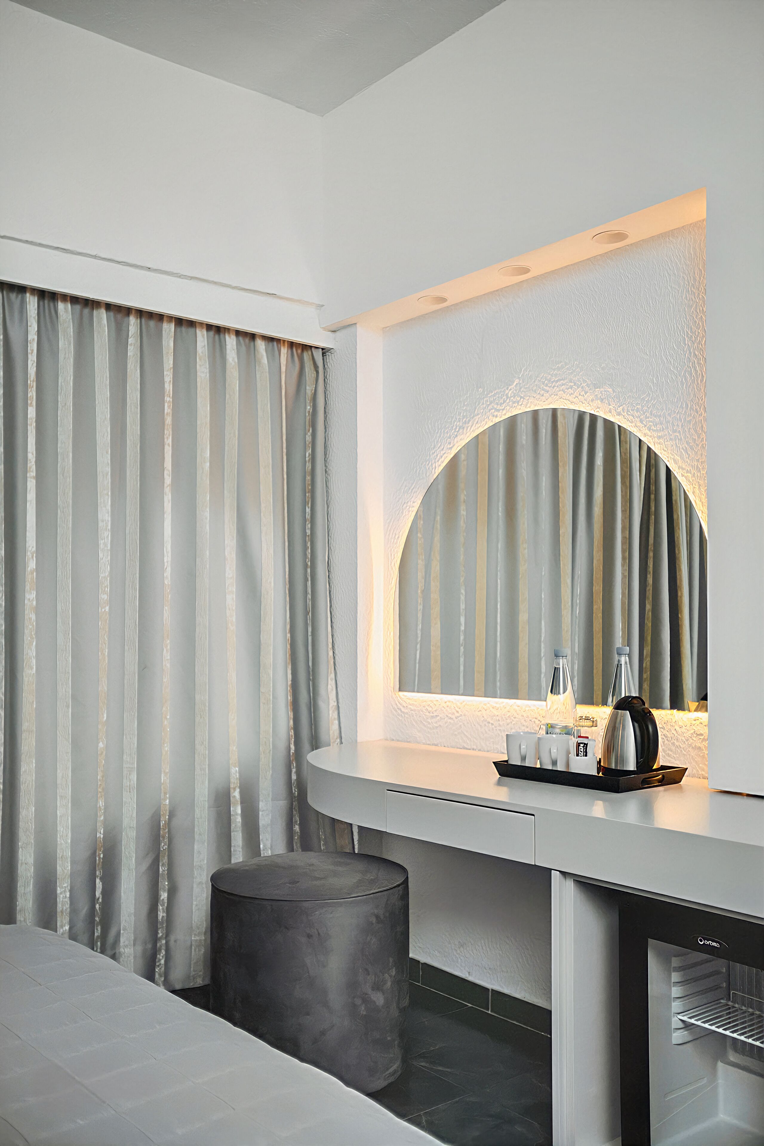 Ein modernes Hotelzimmer mit einer gebogenen Kommode, einem runden Spiegel, einem kleinen runden Tisch mit Wasserkocher und einer Minibar.