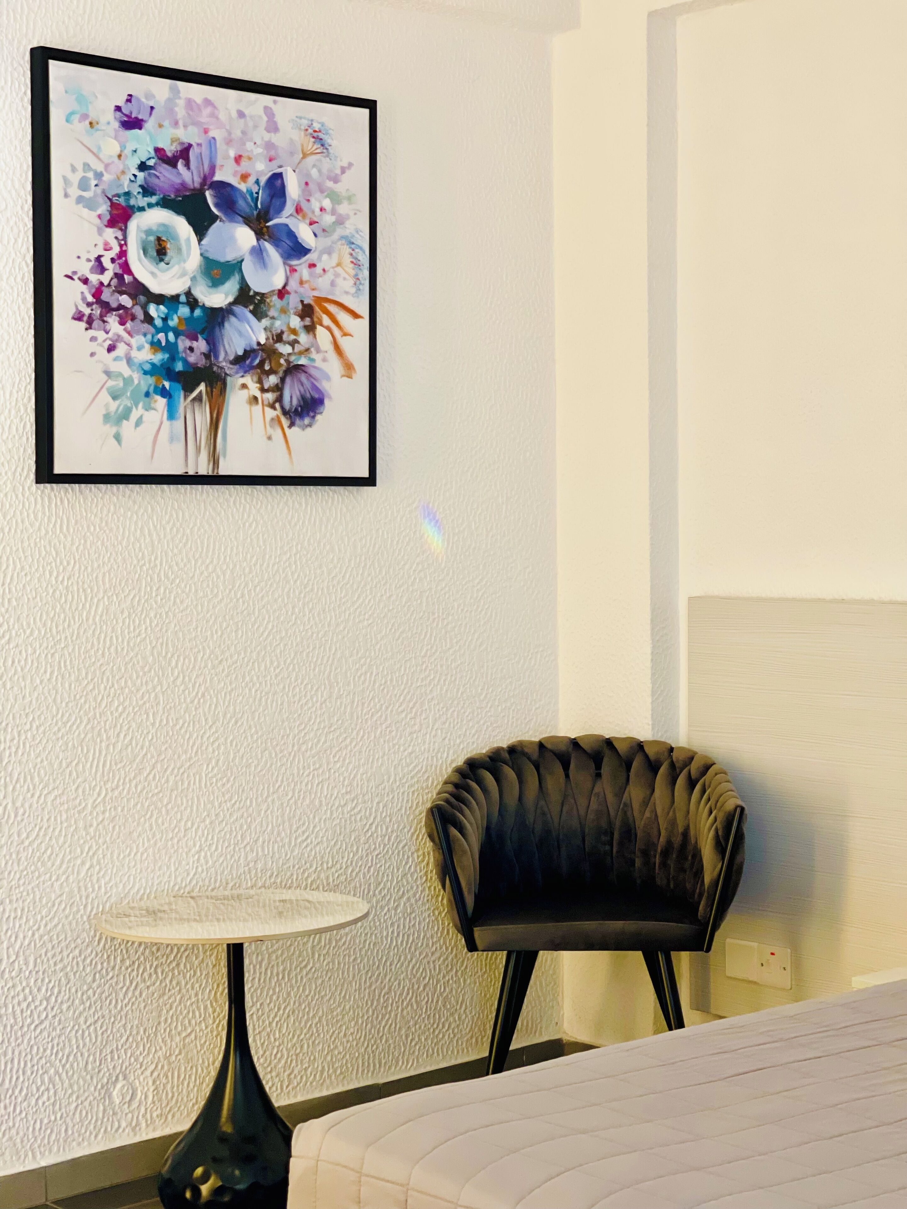 A hotel room with a bed, a small round table, a chair, and a framed floral artwork on the wall.
