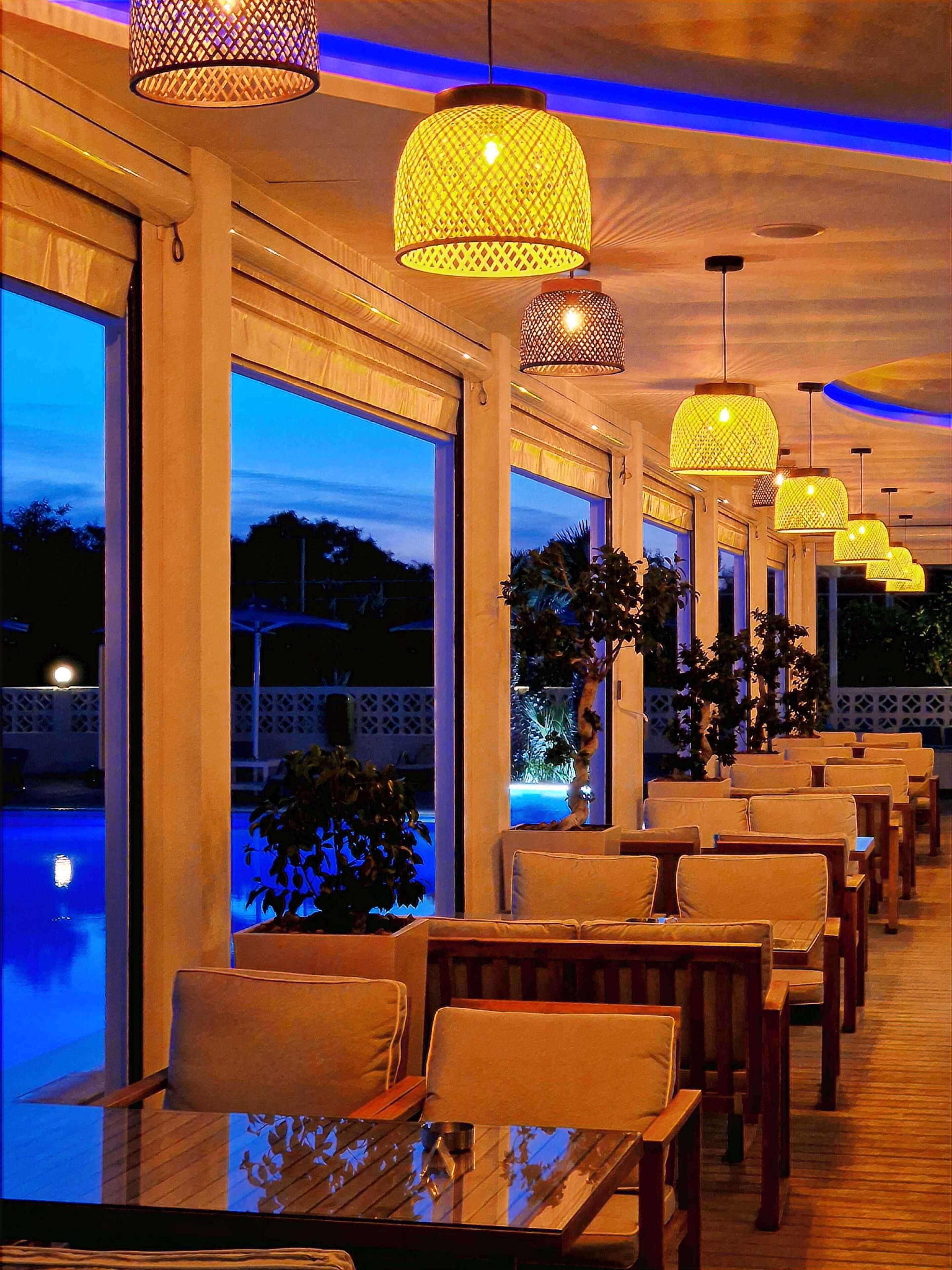 A restaurant with a pool view, featuring multiple tables and chairs, and hanging pendant lights.