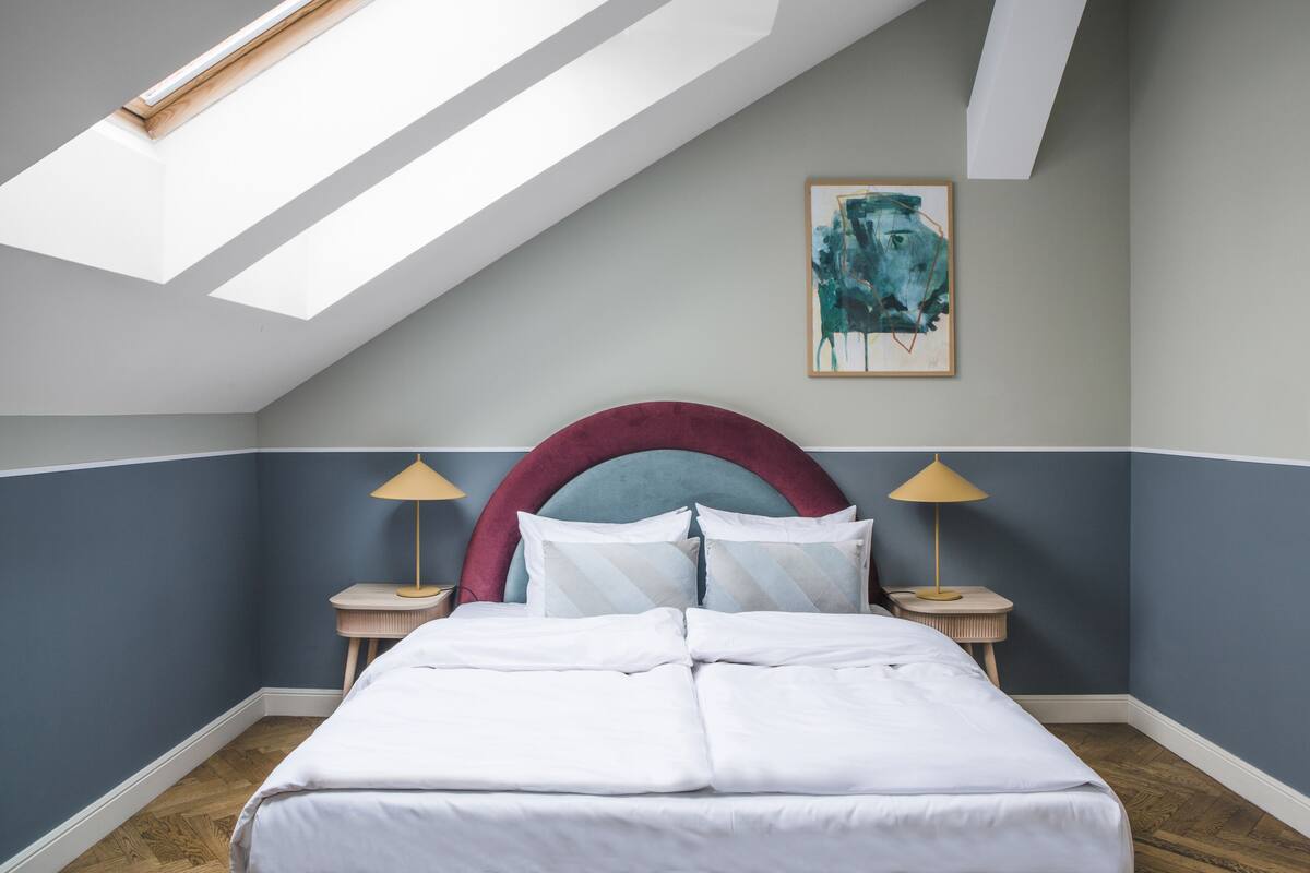 A bedroom with a skylight, a bed with white bedding, a red headboard, and two bedside lamps.