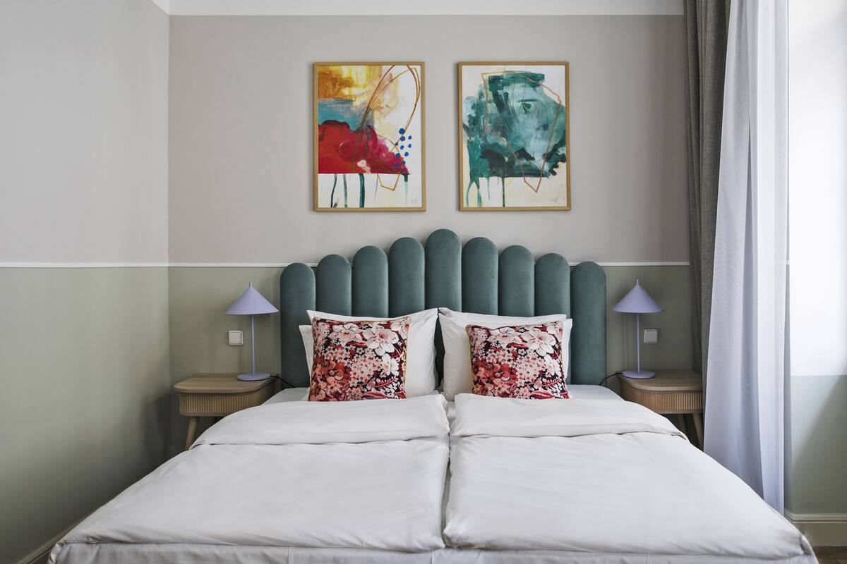 A neatly made bed with white linens and floral pillows, a headboard with a unique design, and two framed artworks on the wall.