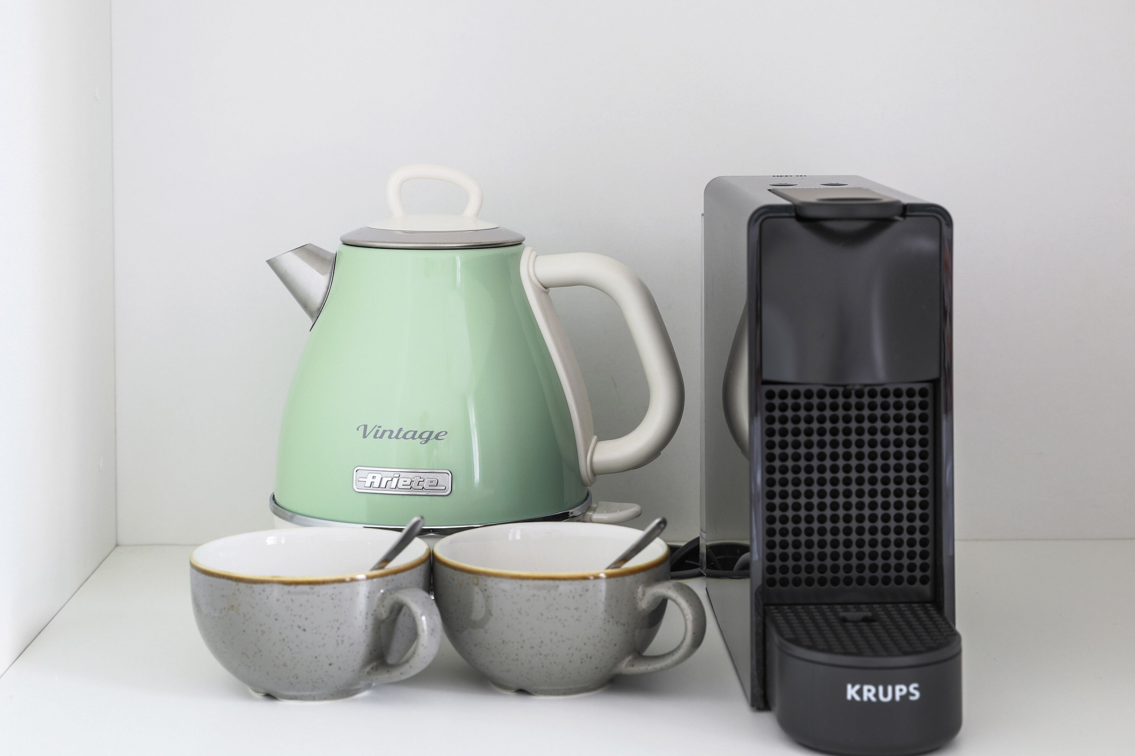 A green kettle, two mugs, and a black Krups electric kettle on a white surface.
