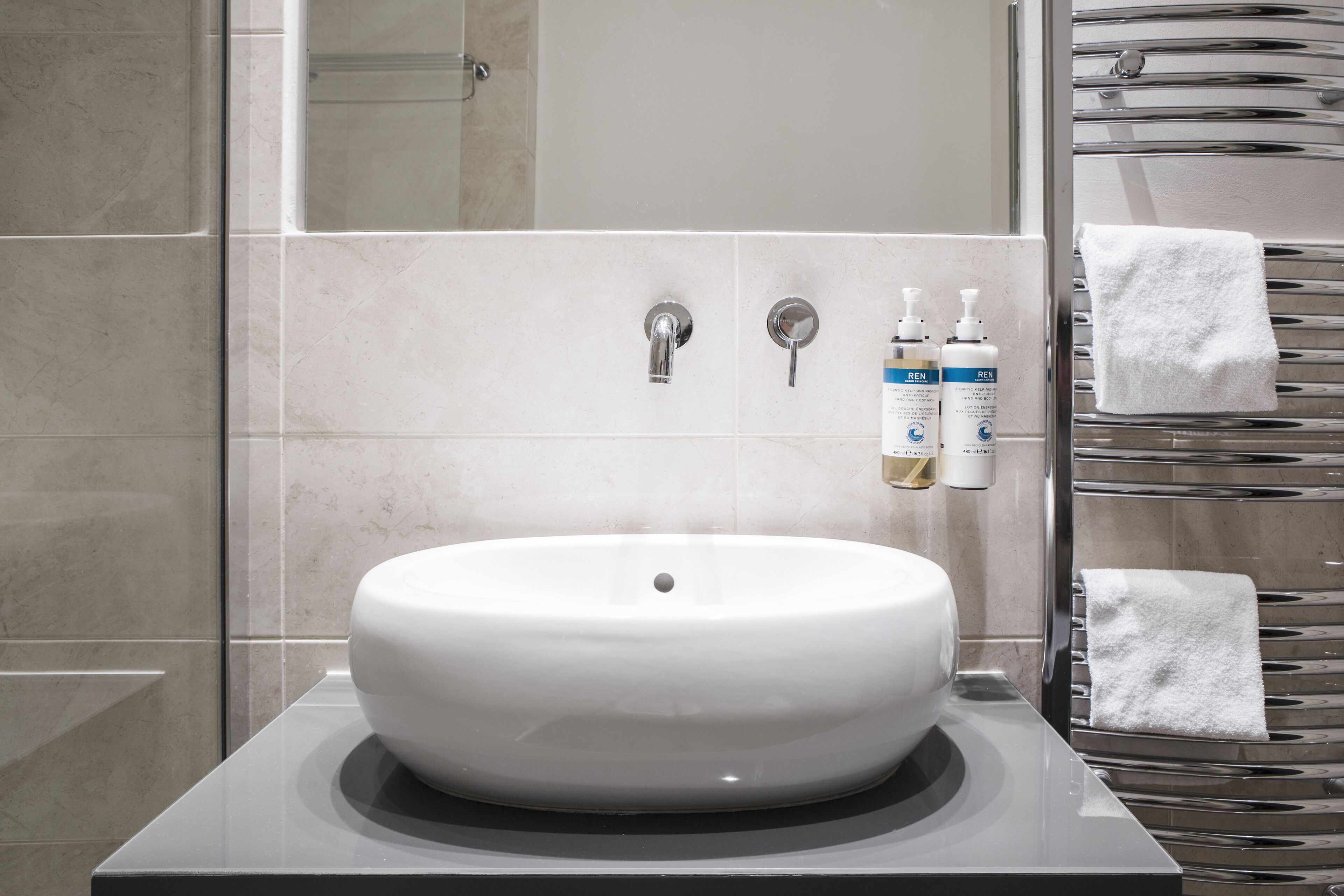A modern bathroom with a large white sink, two faucets, and a towel rack with multiple towels.