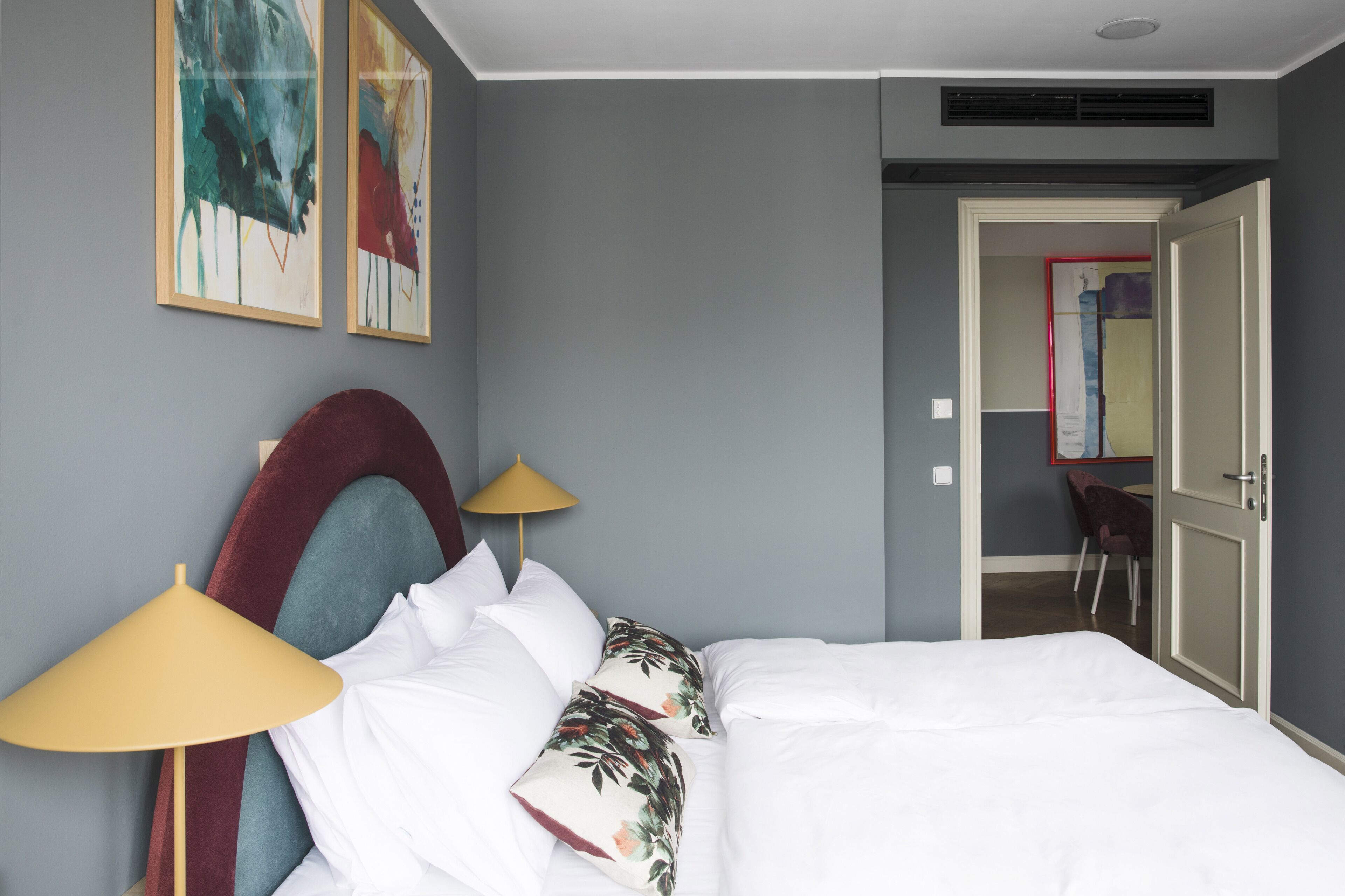 A hotel room with a bed, pillows, a lamp, and artwork on the wall.