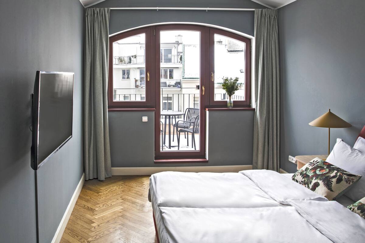 A hotel room with a large bed, a TV, a balcony with a view of buildings, and a small table with a lamp.