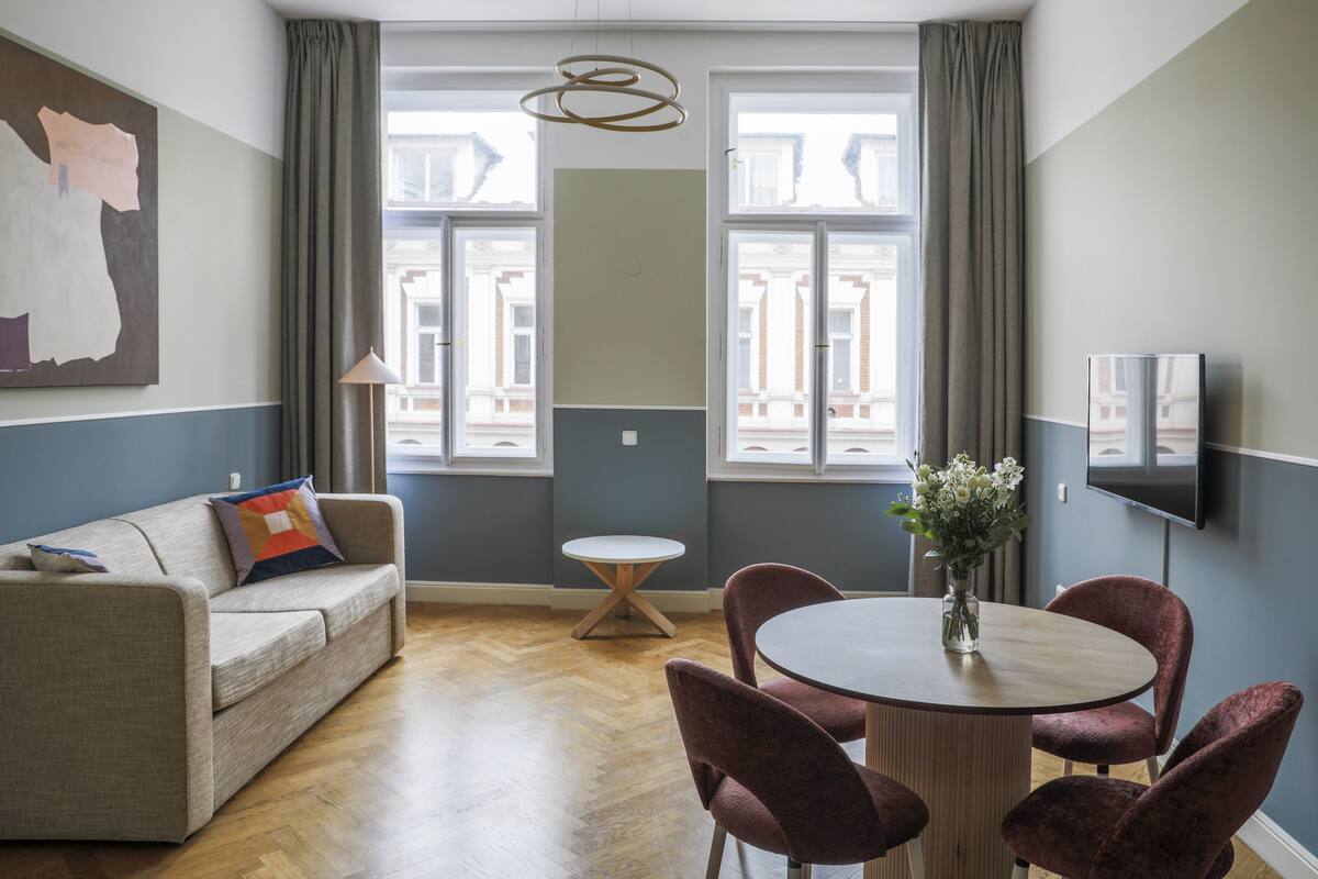 A modern living room with a sofa, a round table with chairs, a vase with flowers, and a flat-screen TV mounted on the wall.