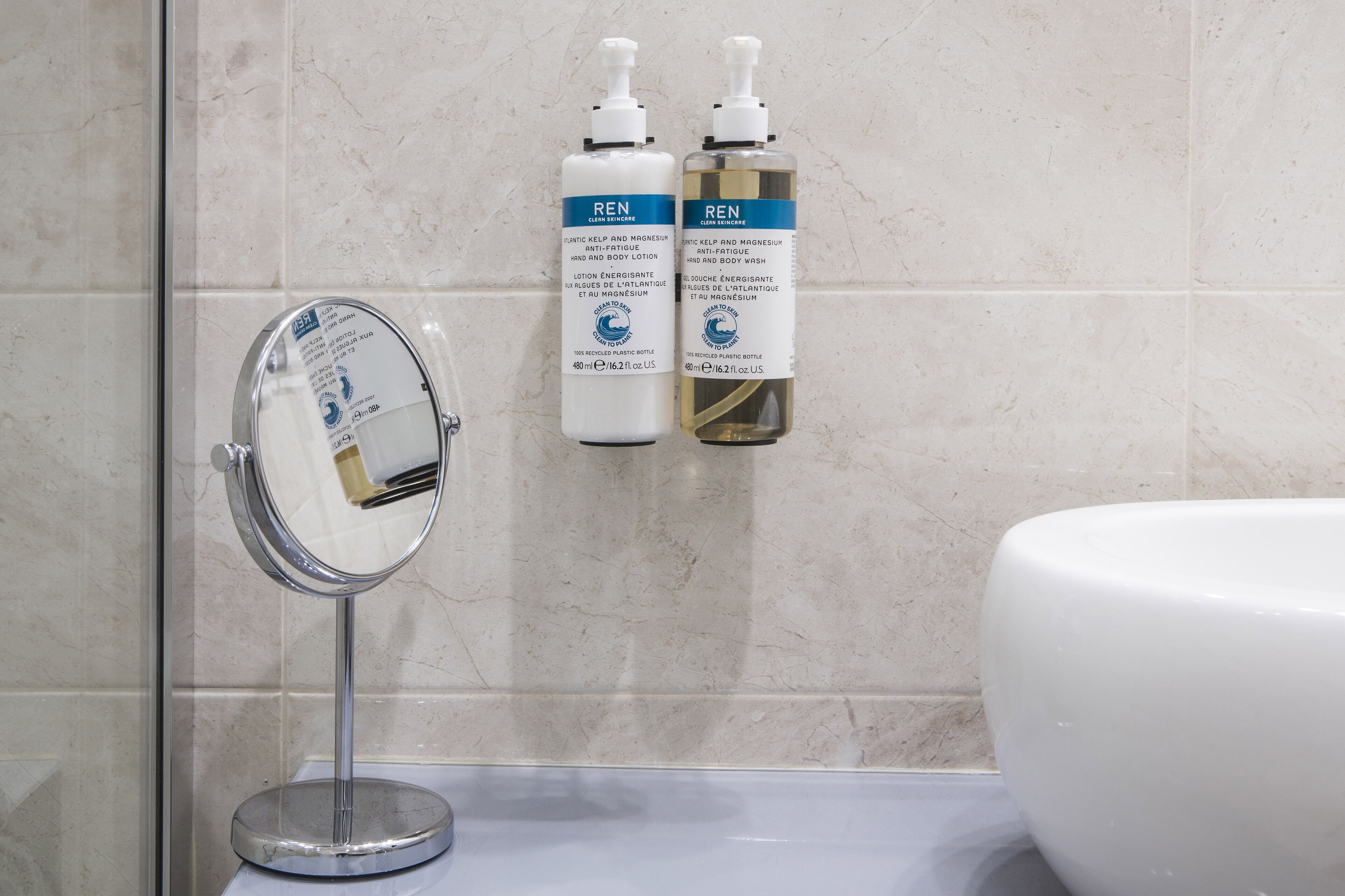 Un baño con un espejo redondo, dos botellas de crema corporal REN y un lavamanos blanco.