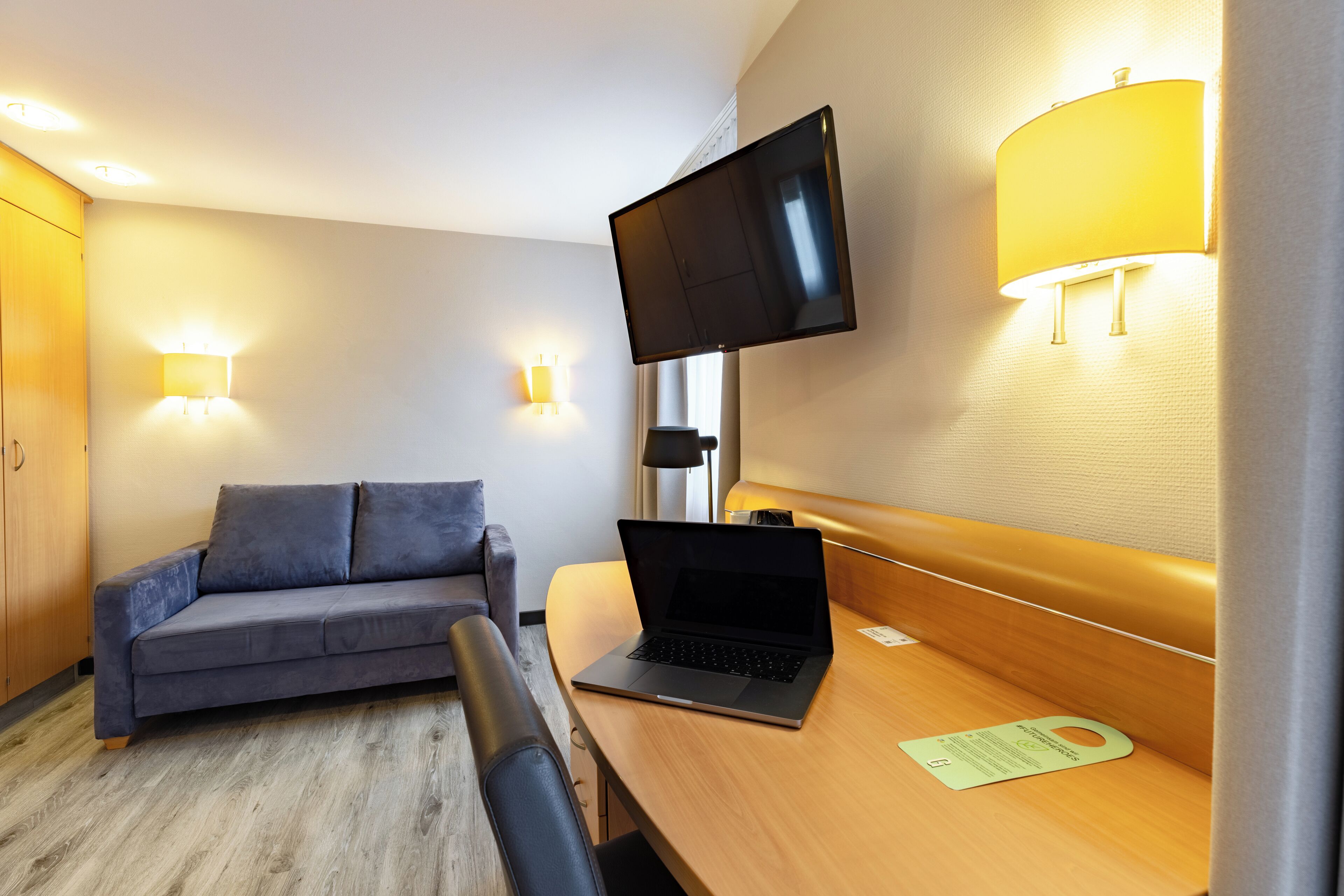Una habitación de hotel moderna con un sofá, un escritorio con una computadora portátil, un televisor de pantalla plana en la pared y dos lámparas de pared.