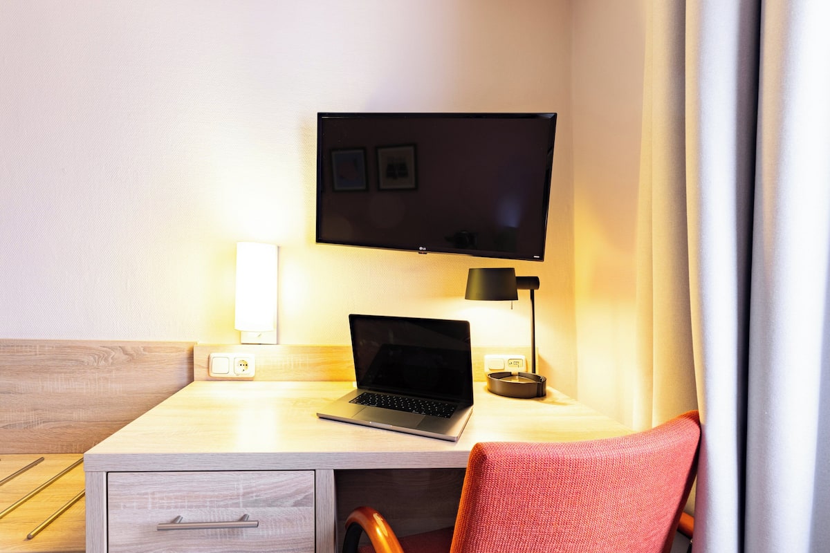 Habitación de hotel con un escritorio de madera, una computadora portátil, un televisor de pantalla plana, una lámpara y una silla roja.