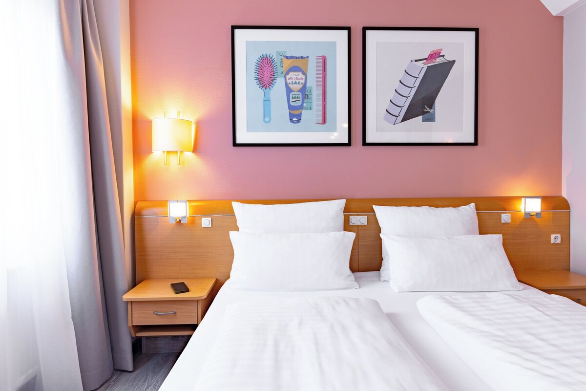 Habitación de hotel con dos camas, una mesita de noche, una lámpara y dos cuadros en la pared.