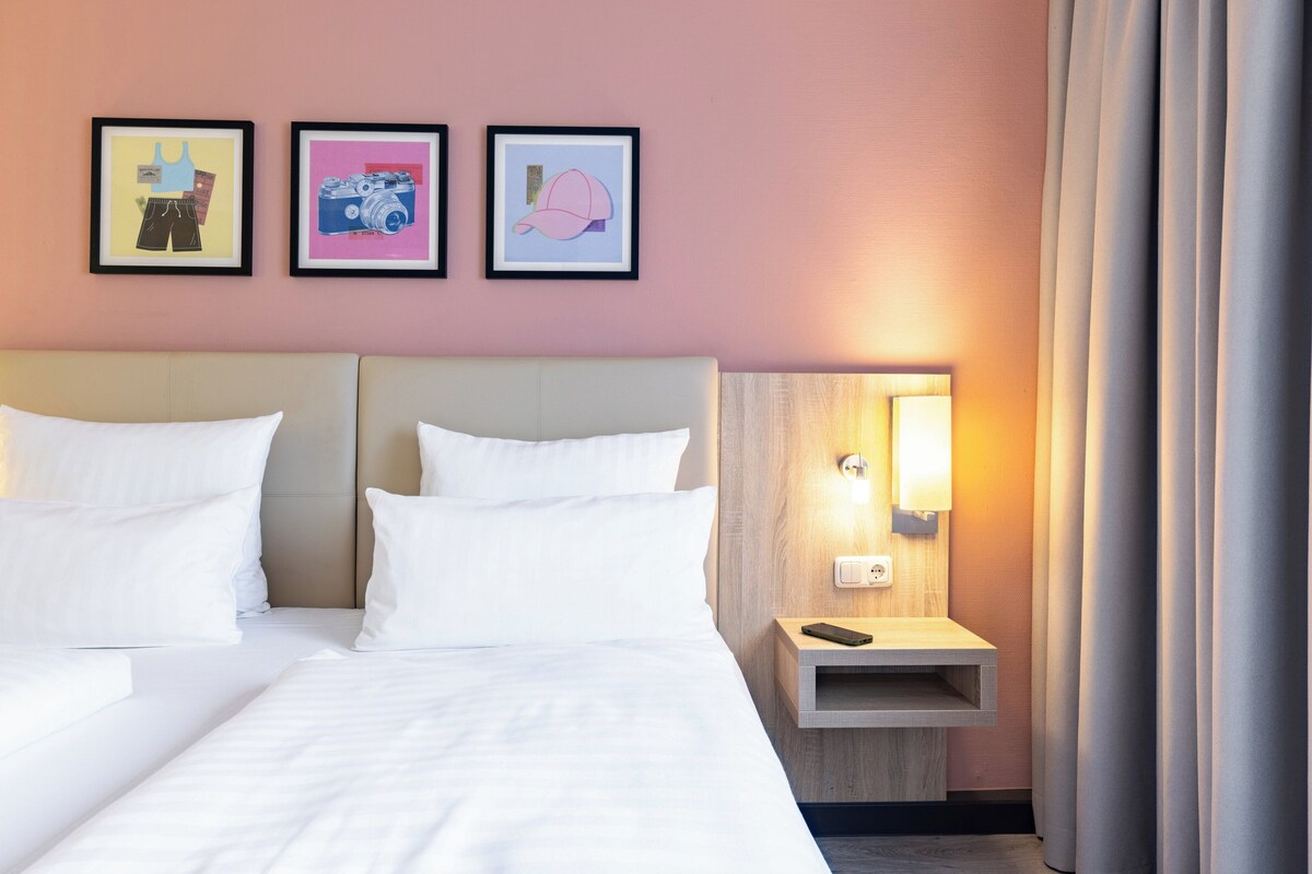 Habitación de hotel con cama, ropa de cama blanca, mesita de noche con lámpara y tres cuadros en la pared.
