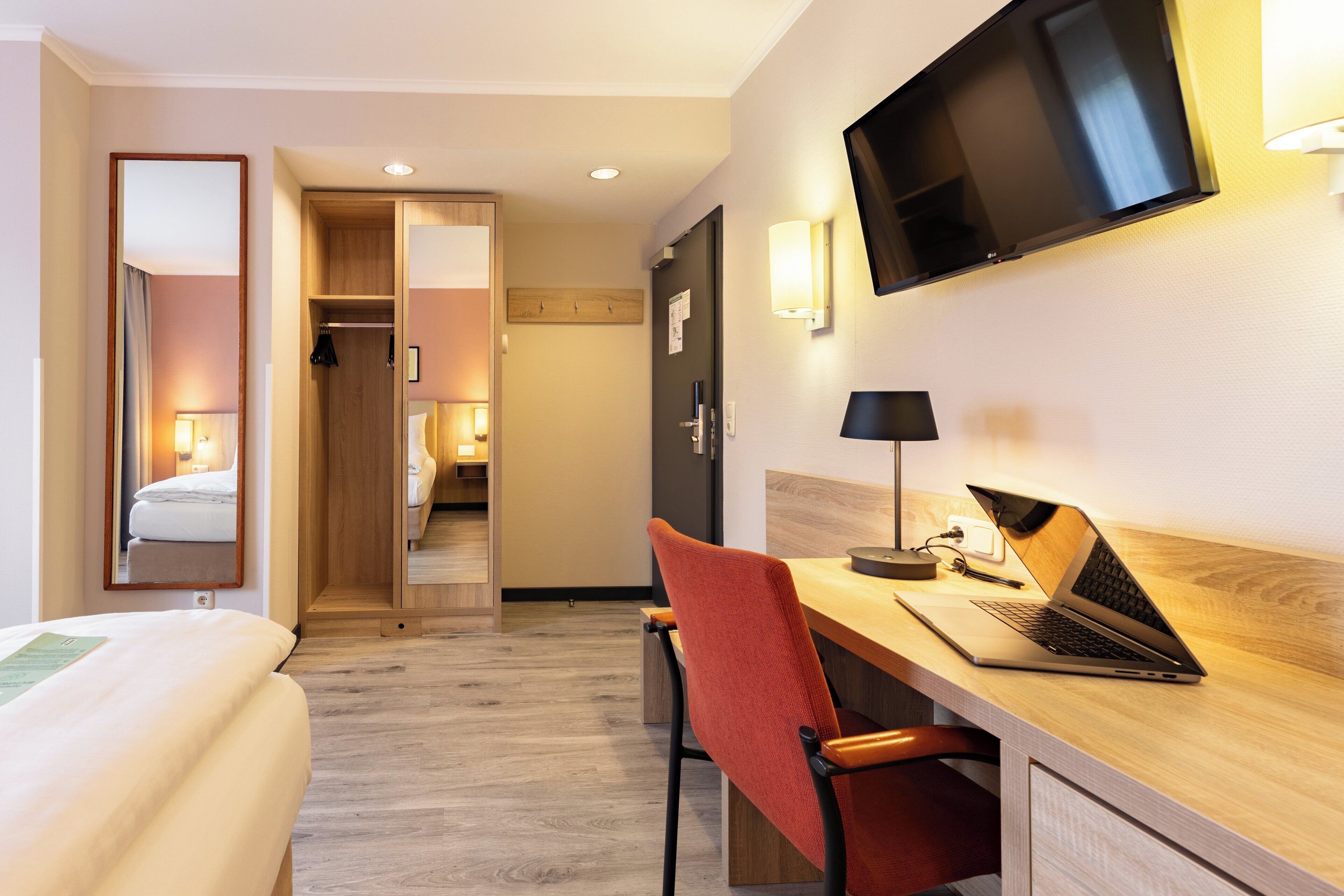 A hotel room with a bed, a desk with a laptop, a TV, and a mirror.