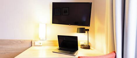 A hotel room with a wooden desk, a laptop, a flat-screen TV, a lamp, and a red chair.