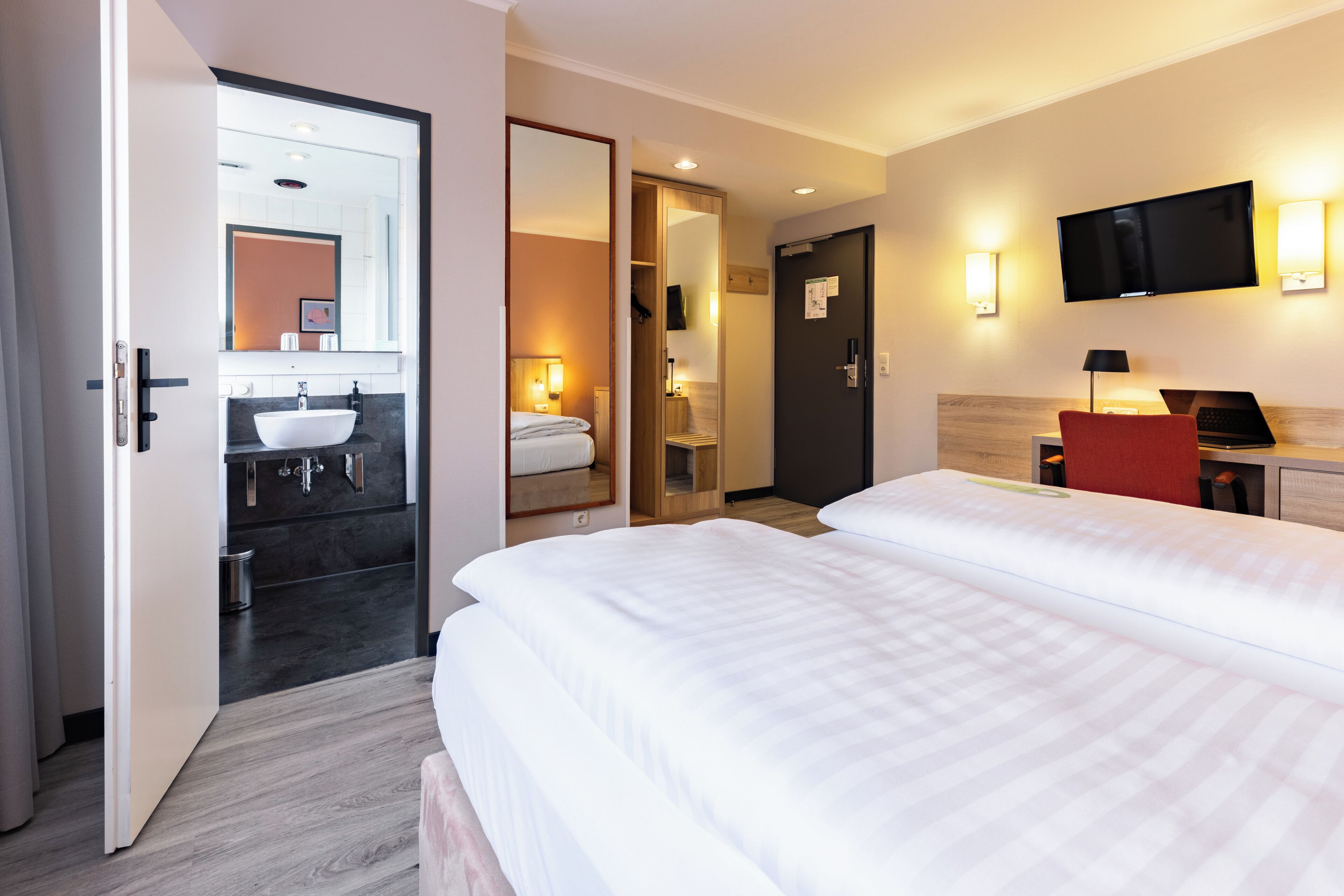 Una habitación de hotel con una cama grande, una mesita de noche con lámpara, un televisor montado en la pared y un baño con lavamanos y espejo.