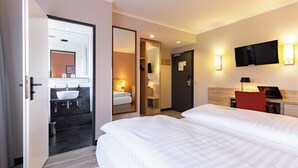 Una habitación de hotel con una cama grande, una mesita de noche con lámpara, un televisor montado en la pared y un baño con lavabo y espejo.