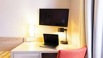 Une chambre d’hôtel avec un bureau en bois, un ordinateur portable, un téléviseur à écran plat fixé au mur, une chaise rouge et une table de chevet avec une lampe.
