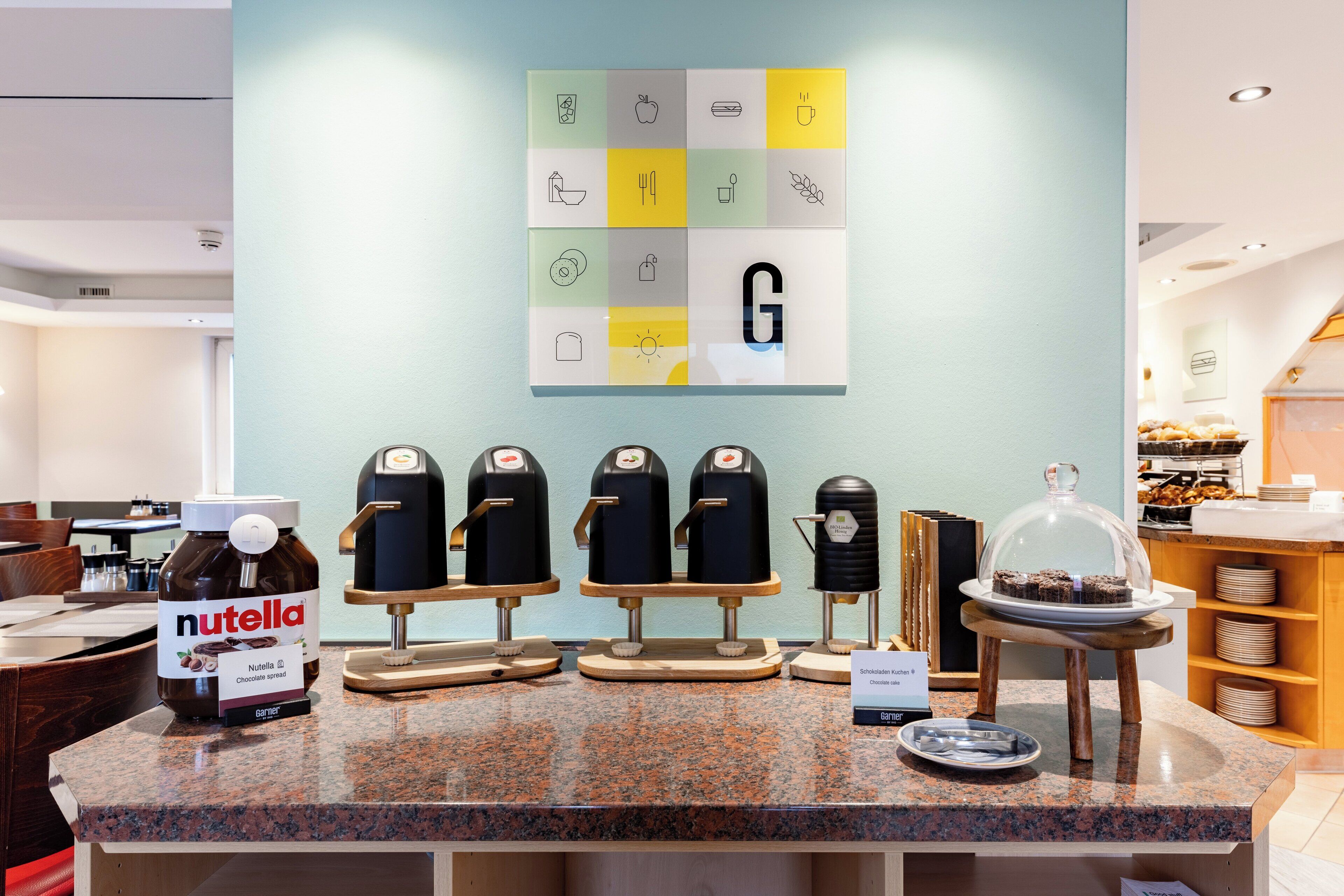A coffee station with Nutella and coffee machines, a display of food items, and a wall with a letter 'G'.