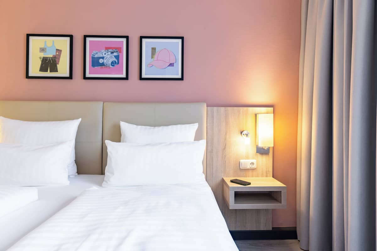 Habitación de hotel con cama, ropa de cama blanca, mesita de noche con lámpara y tres cuadros en la pared.