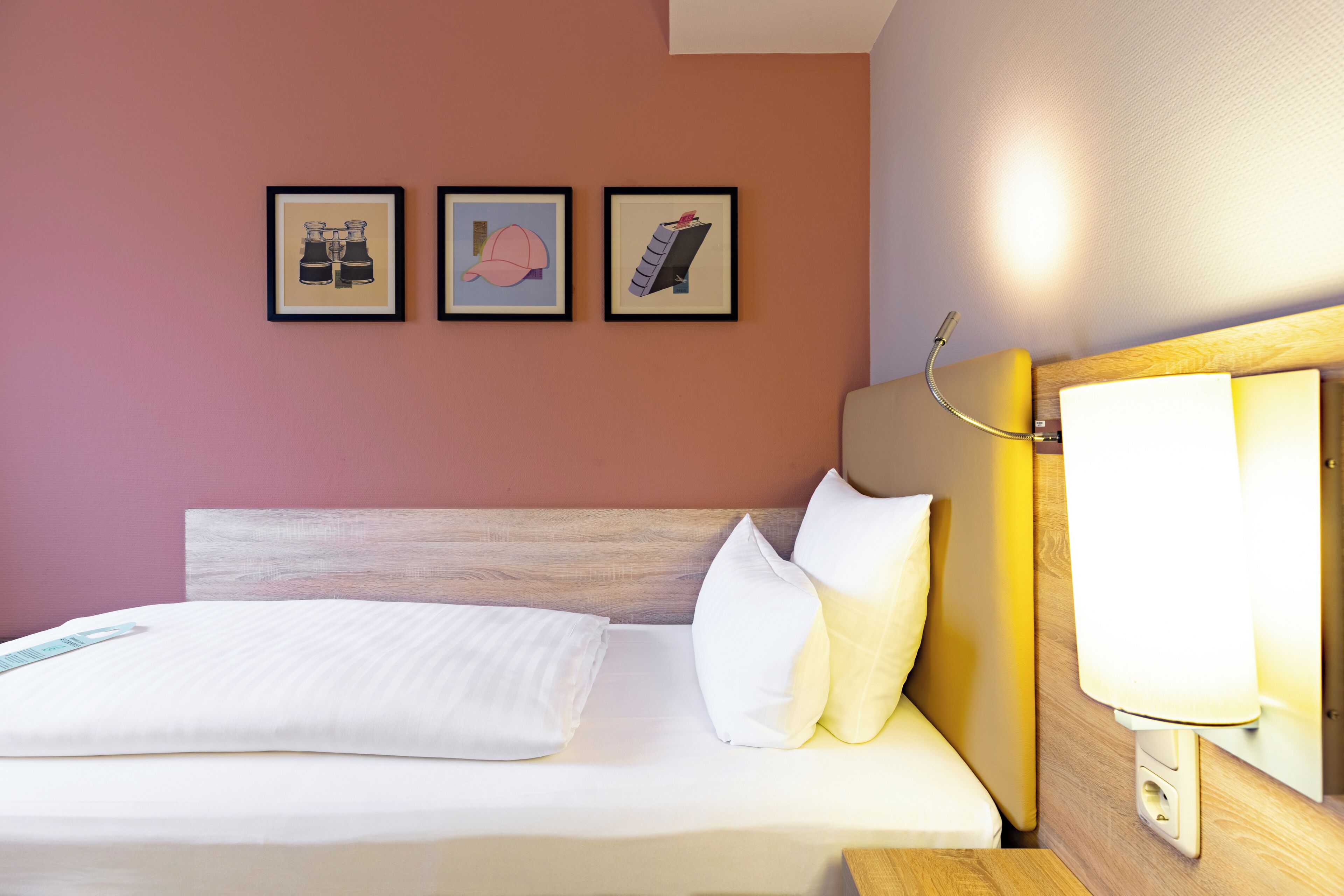 Habitació d'hotel amb un llit, roba de llit blanca, un panell de capçalera de fusta, una tauleta de nit amb una làmpada i tres quadres al mur.