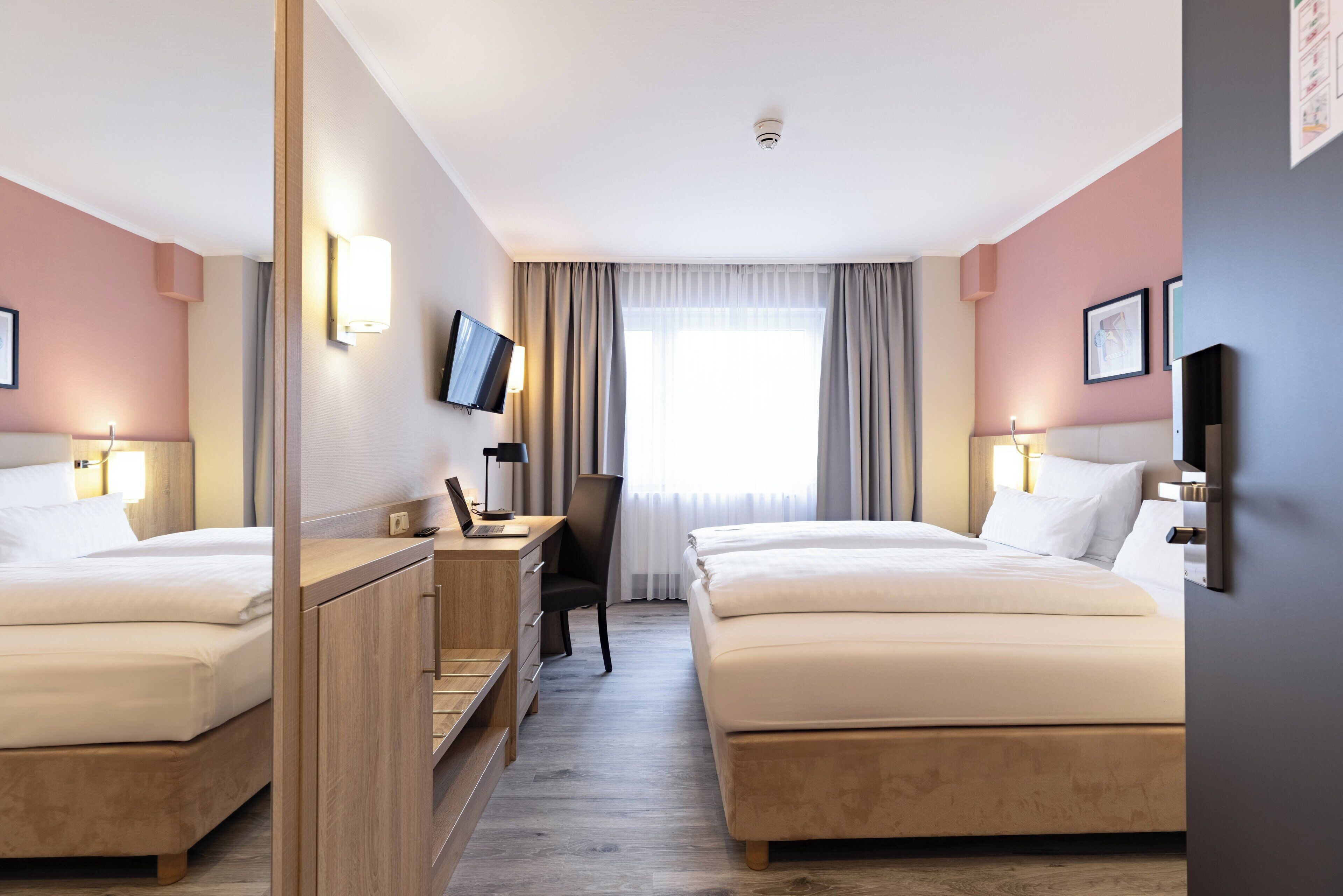 Habitación de hotel con dos camas, un escritorio con una computadora portátil, una silla, un televisor montado en la pared y un ventanal con cortinas.