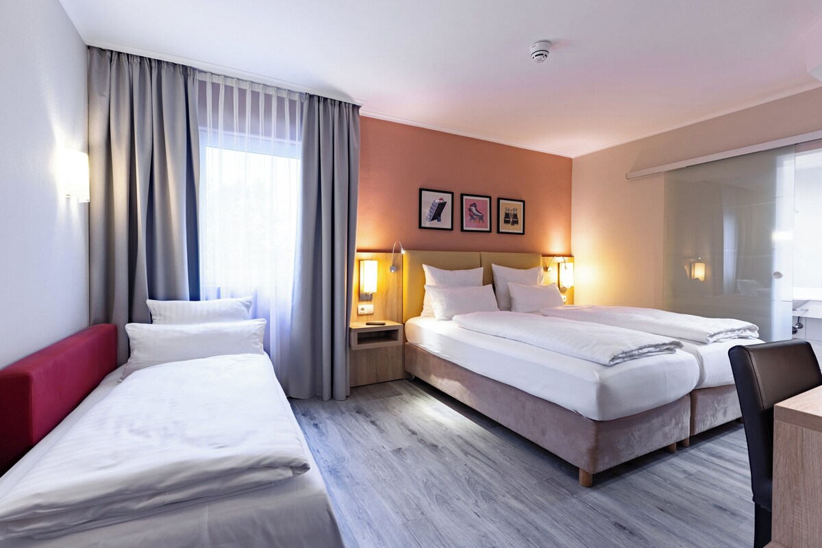 Una habitación de hotel con dos camas, cabecera roja, suelo de madera y un ventanal con cortinas.