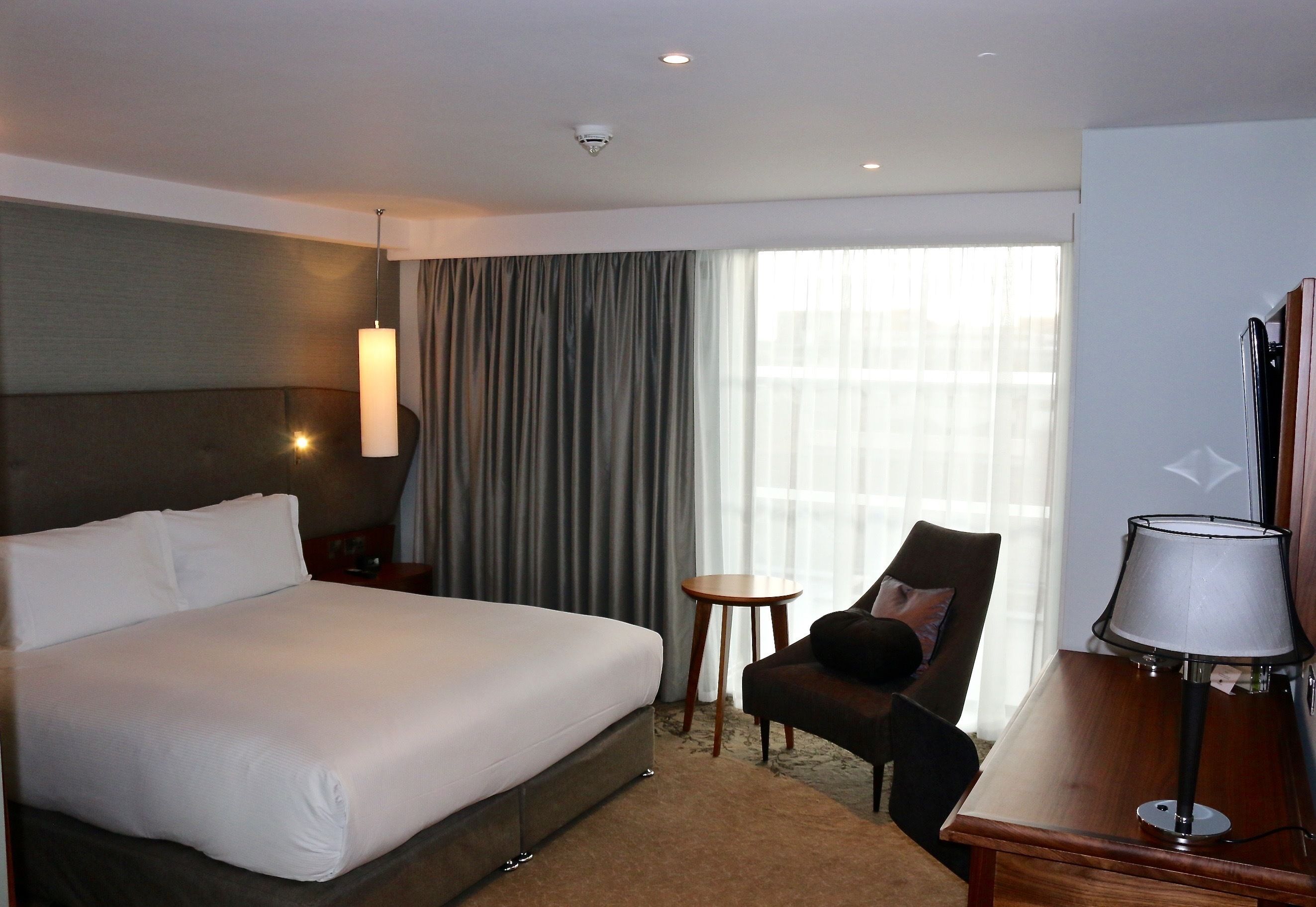 Habitación de hotel con una cama grande, un escritorio, una silla, una lámpara y vistas al exterior por la ventana.