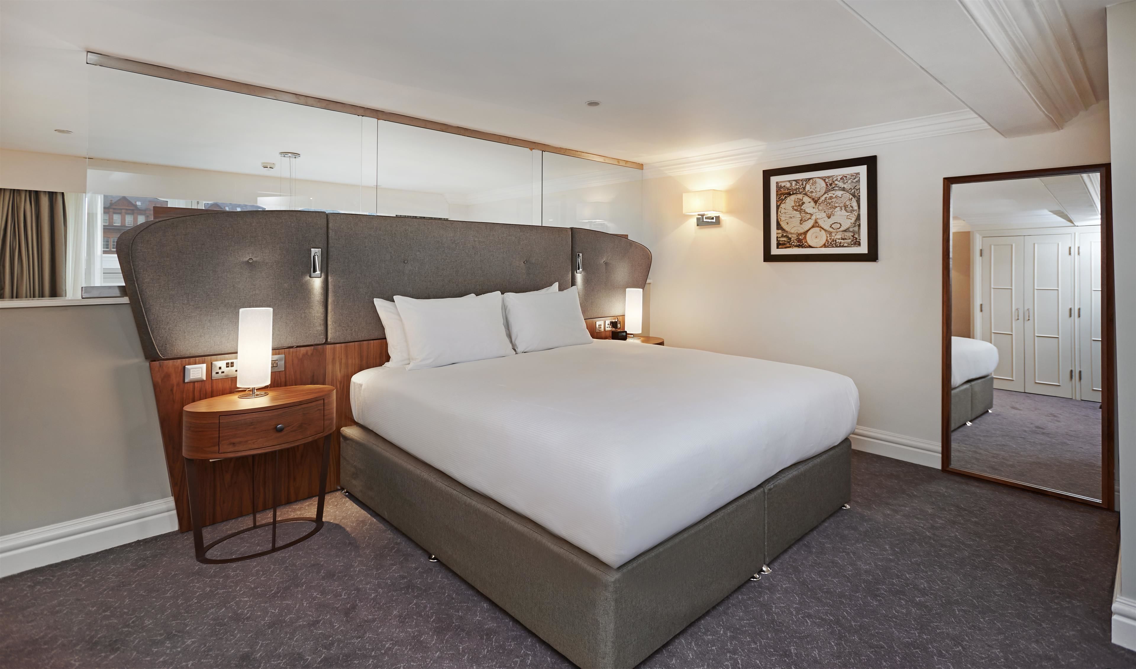 Una habitación de hotel con una cama grande, una mesita de noche con lámpara, una luz en la pared, un cuadro enmarcado y un espejo.