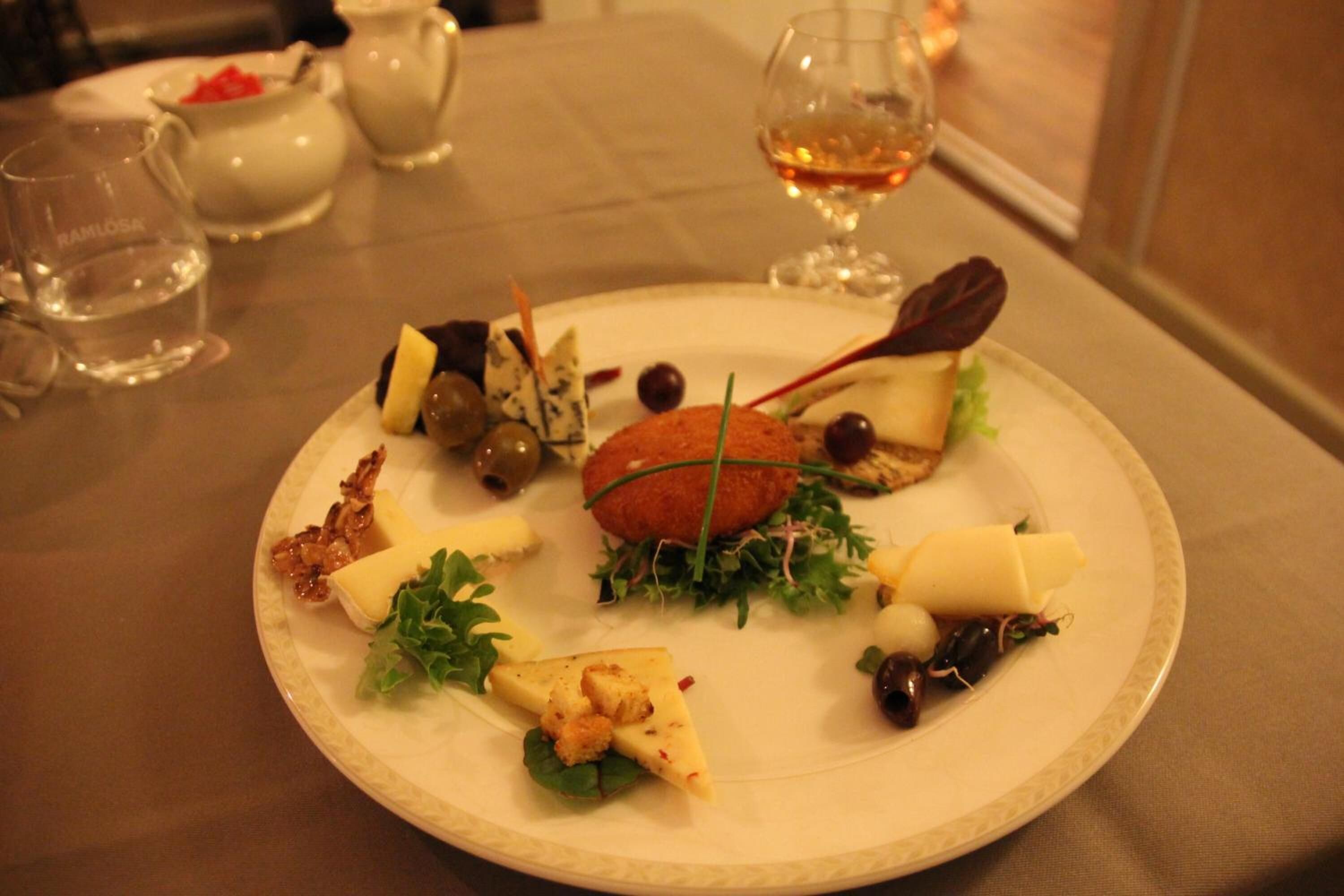 Un plato de aperitivos variados, con una croqueta, queso, aceitunas y vegetales, junto con una copa de vino y un vaso de agua al fondo.
