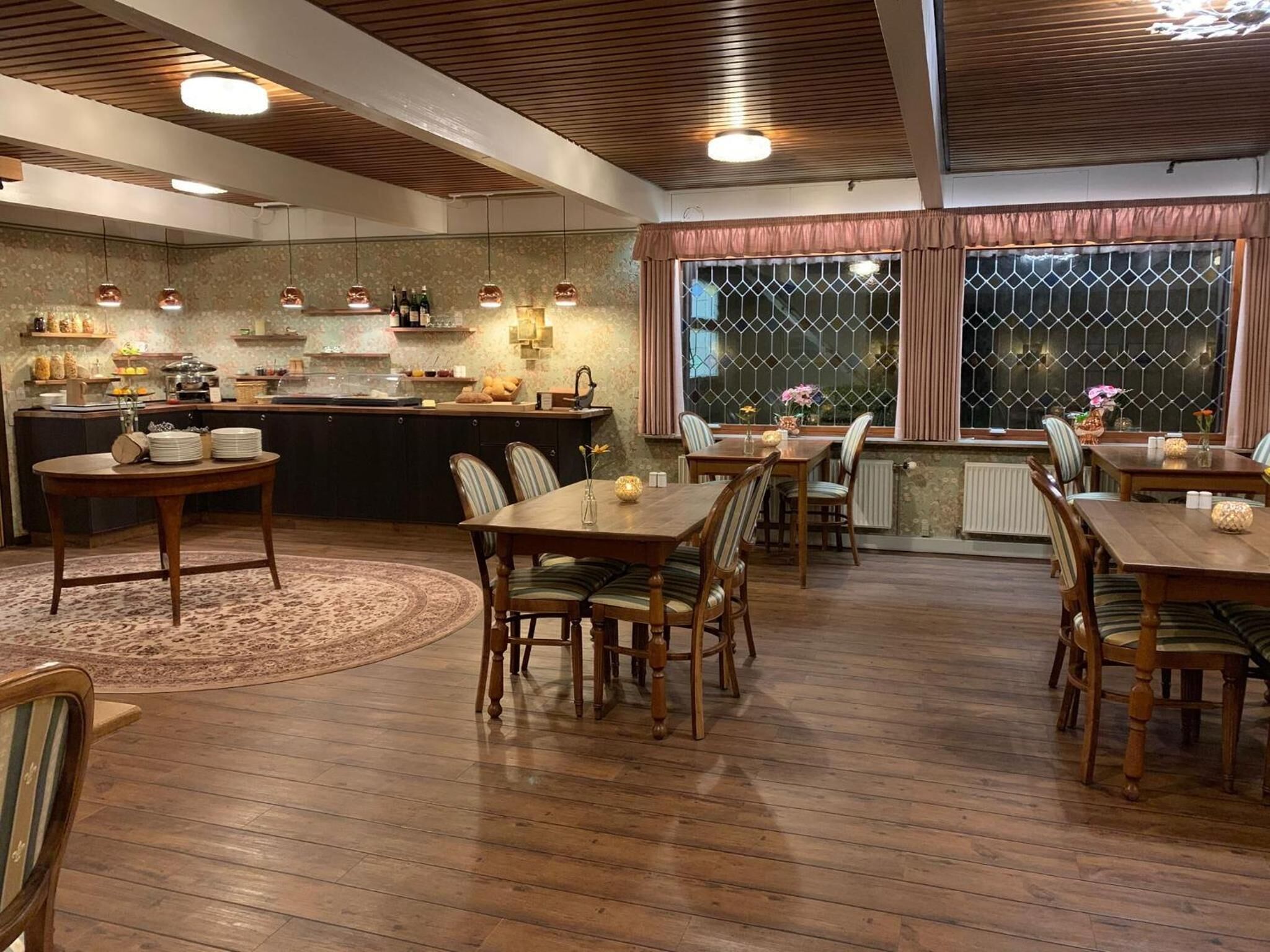 A spacious dining area with wooden tables and chairs, a kitchen in the background, and a large window with curtains.