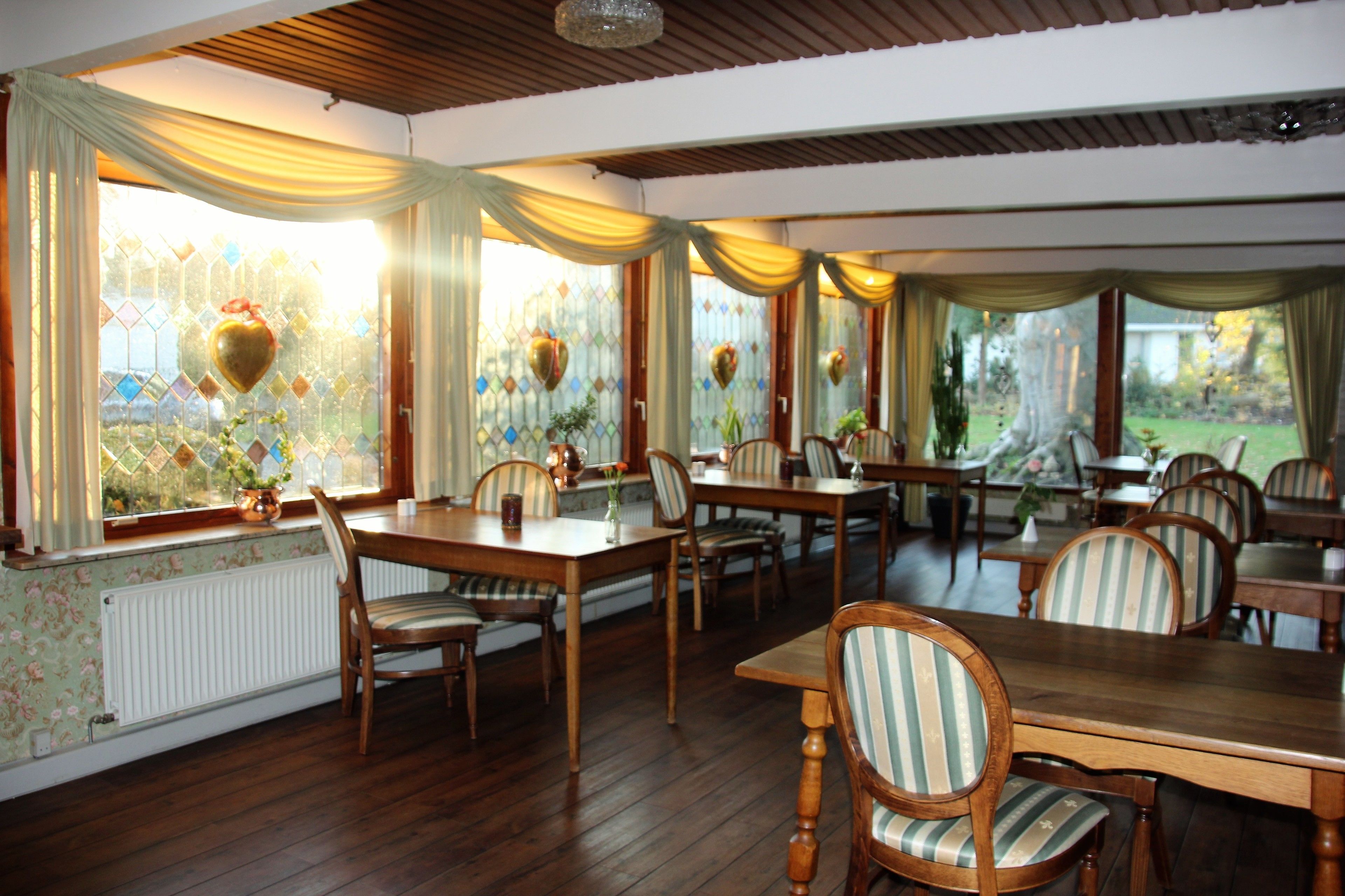 A dining area with wooden tables and striped chairs, large windows with curtains, and a view of a garden.