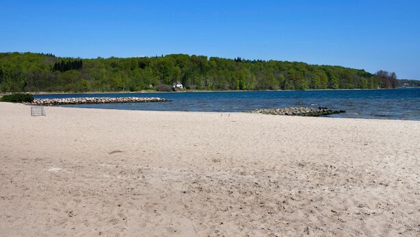 A sandy beach with a rocky shoreline and a forested hill in the background. - Bov Kro (Padborg)