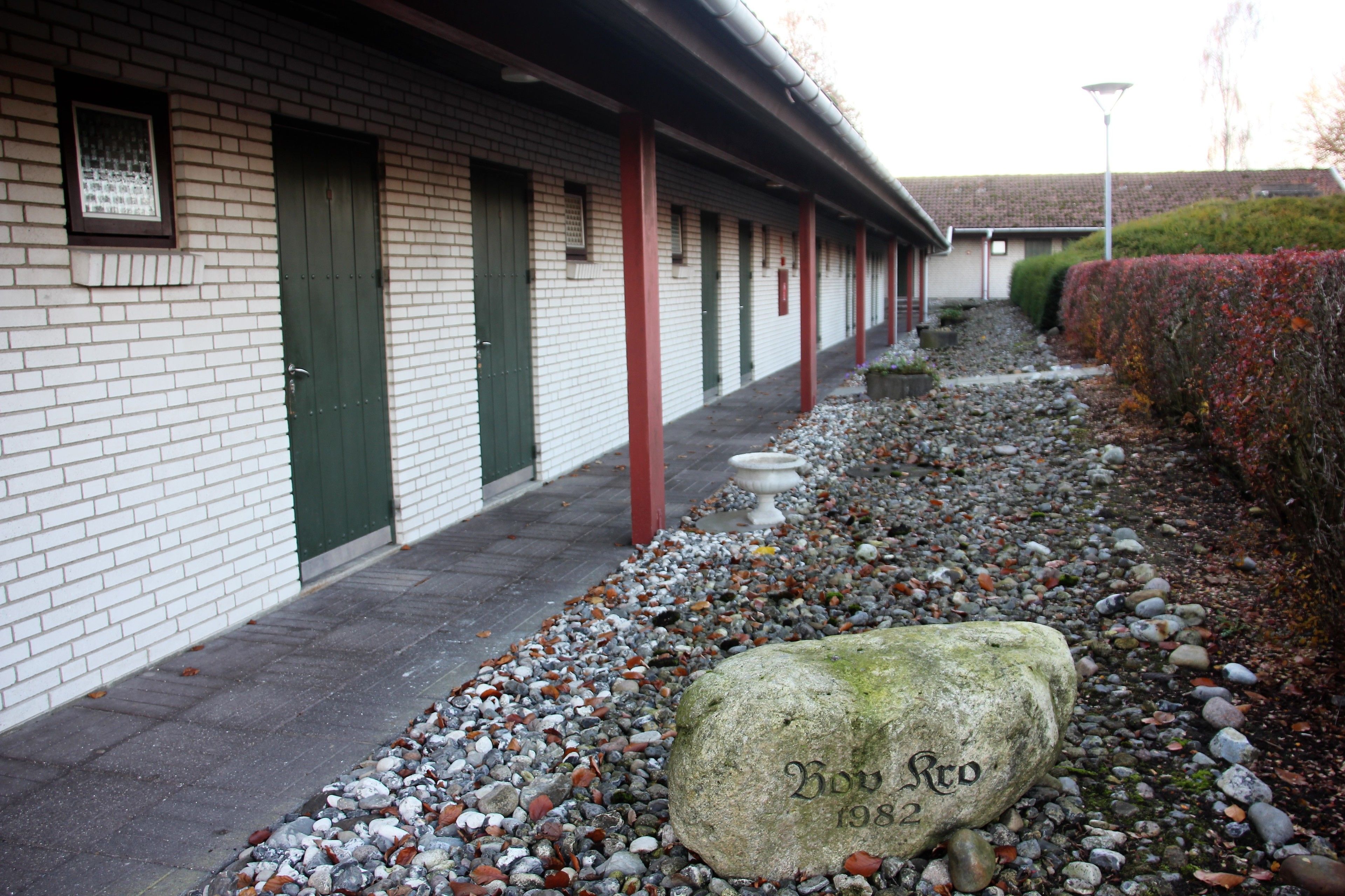 A building with a brick facade, green doors, and a pathway lined with pebbles and a large rock with 'Bow Rd 1982' inscribed.