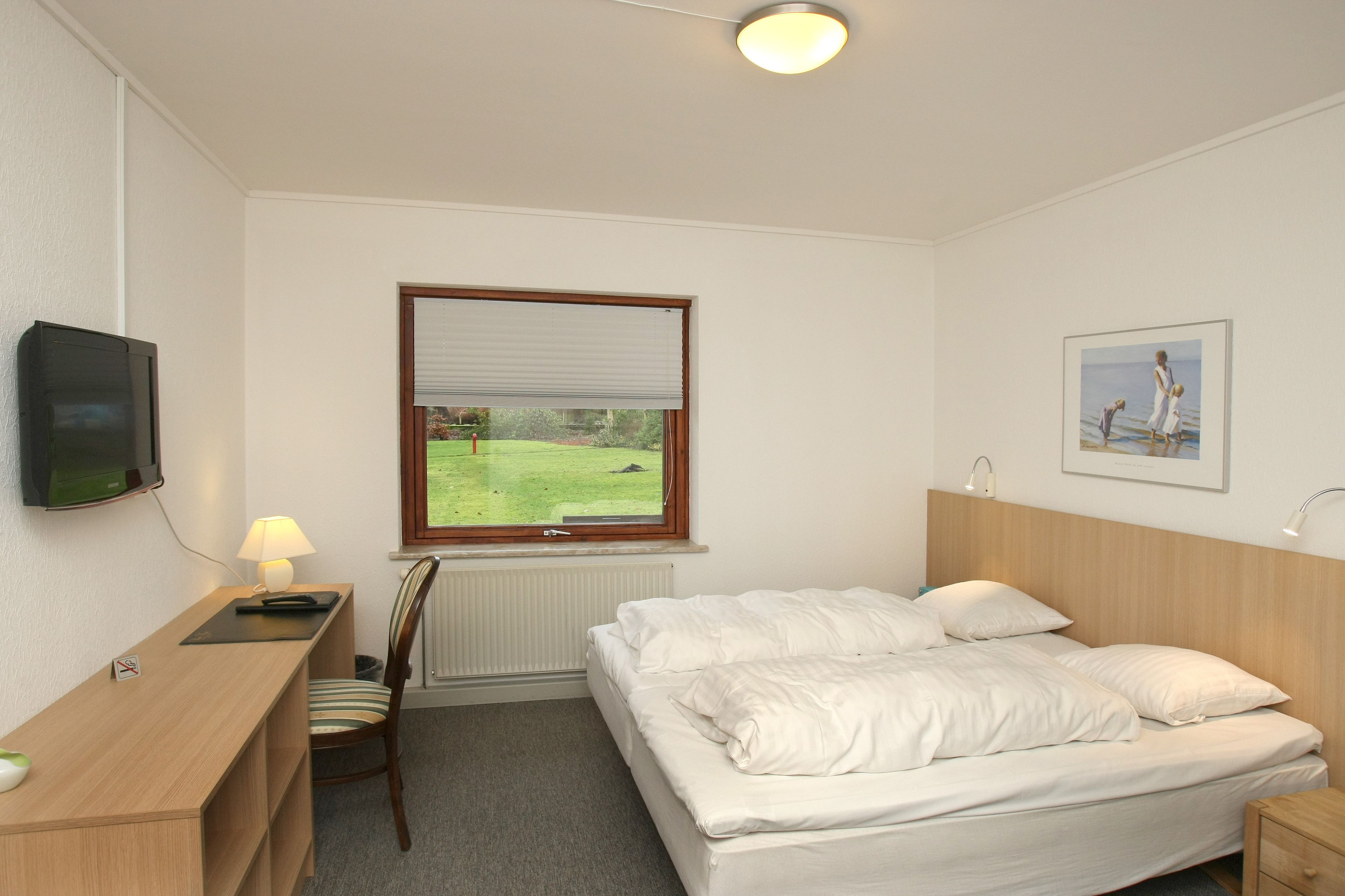Hotelkamer met een bed, een bureau met lamp, een stoel, een televisie, een raam met uitzicht op een groen veld en een lijst met een afbeelding aan de muur.