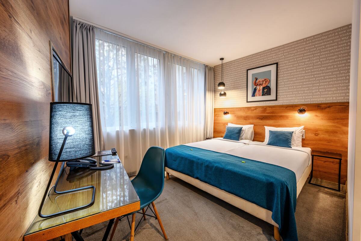 A hotel room with a wooden headboard, a desk with a lamp, a bed with blue bedding, and a framed picture on the wall.