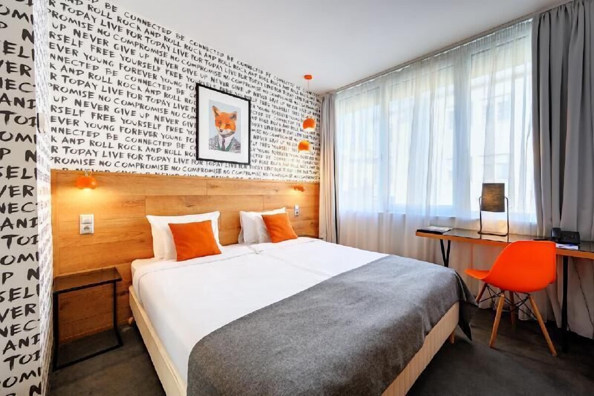 A hotel room with a large bed, a desk, and a chair. There is a wall with text and a framed picture.