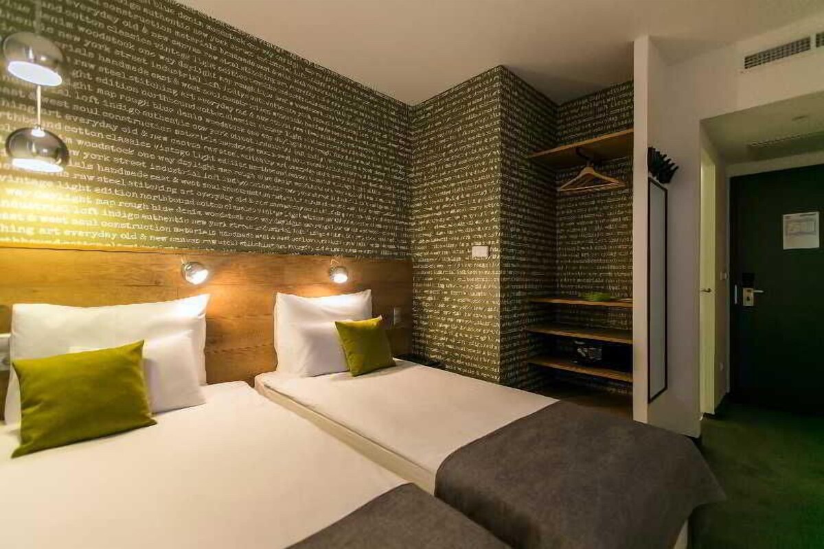 A hotel room with two beds, a wooden headboard, a green wall with text, and a closet with hanging clothes.